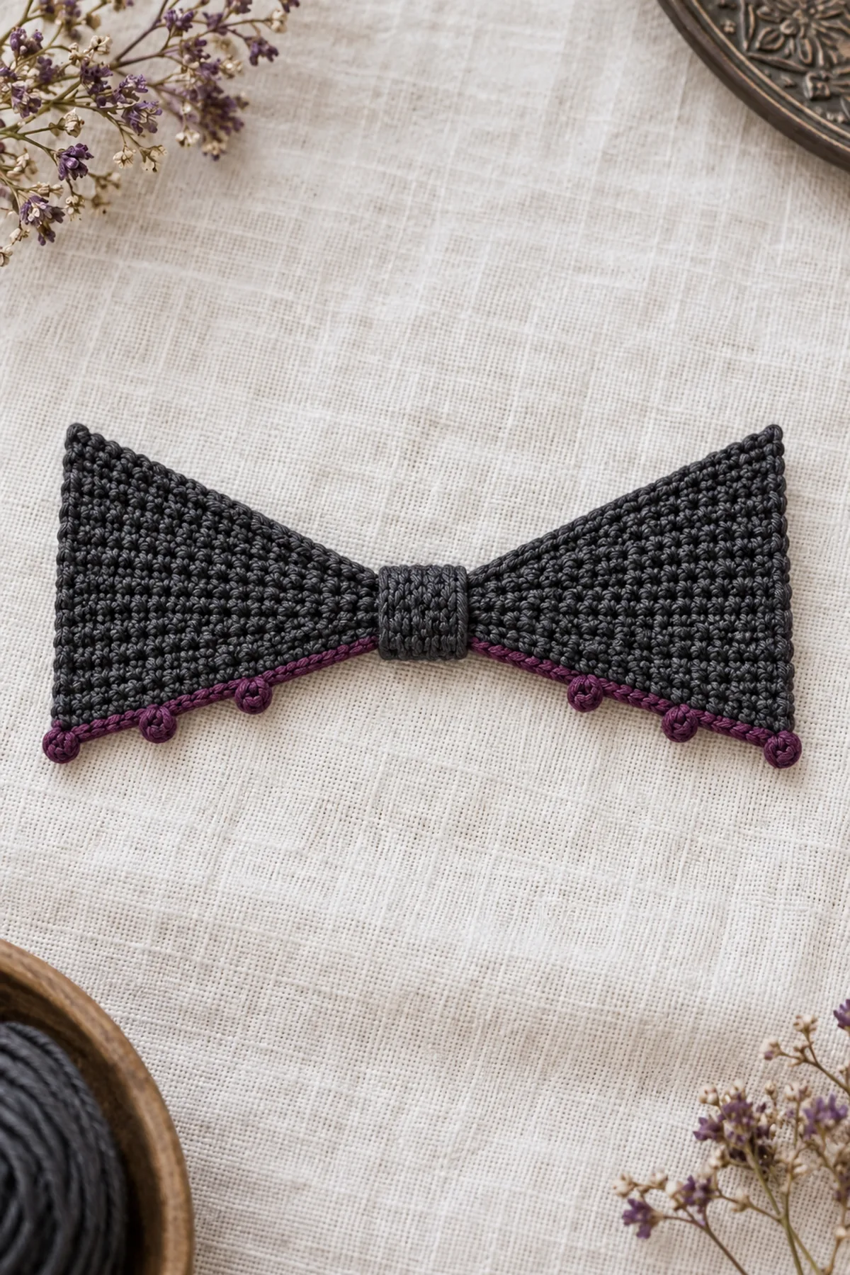One original adult crochet bat-wing bow tie on pale ivory linen: two symmetrical flat charcoal cotton triangular wedges narrow at center and broad at outer ends, straight upper slopes, lower slopes edged in plum with exactly three tiny picot points per wing, narrow charcoal center wrap, about 4.3 inches wide and 2 inches tall. No stuffed bat body, no eyes, no ears, no head, no neck cord, no large scallops
