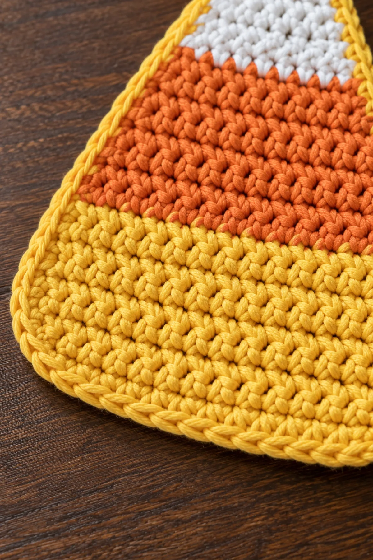 Macro close crop of a flat single-layer candy corn crochet coaster showing yellow single-crochet edging smoothly rounding broad base corner, yellow body stitches and orange stripe visible, no raised thick seams