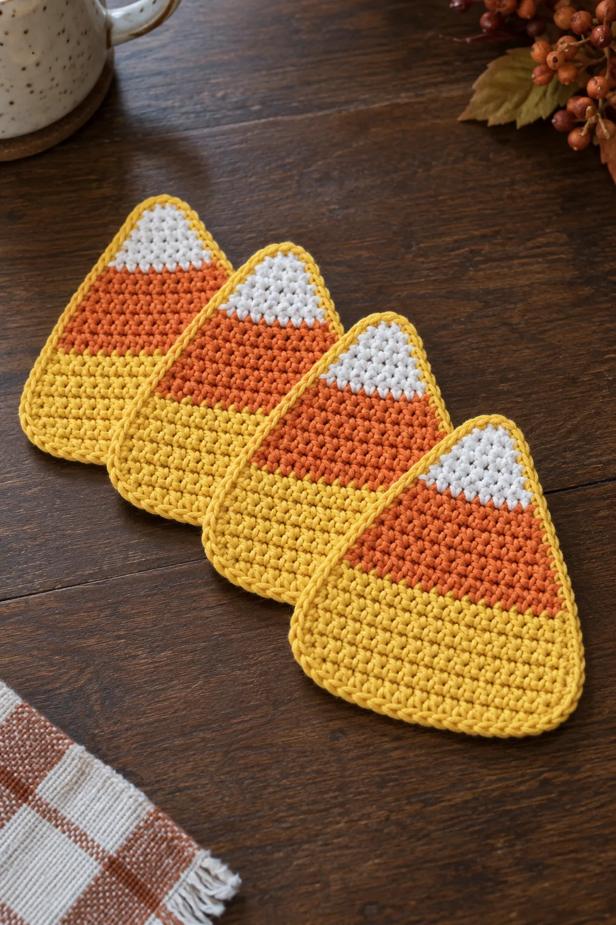 Exactly four flat single-layer cotton crochet candy corn coasters fanned on a dark walnut tabletop, each a broad rounded triangle with small white tip, narrow orange center stripe and wide golden-yellow base, thin yellow single-crochet border around entire shape, no stuffing, simple coarse single-crochet texture
