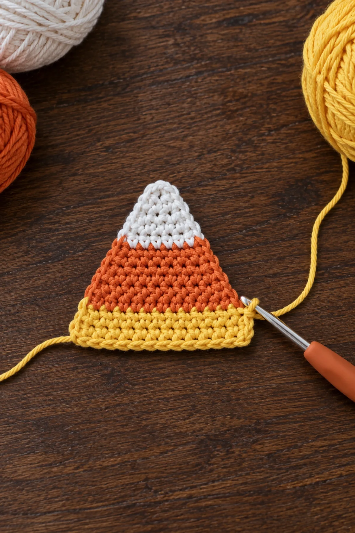 One small flat crochet triangle on hook, white rounded tip and narrow orange stripe completed, two yellow rows just started along wider working edge, no border yet; photographed top down on dark walnut, not stuffed