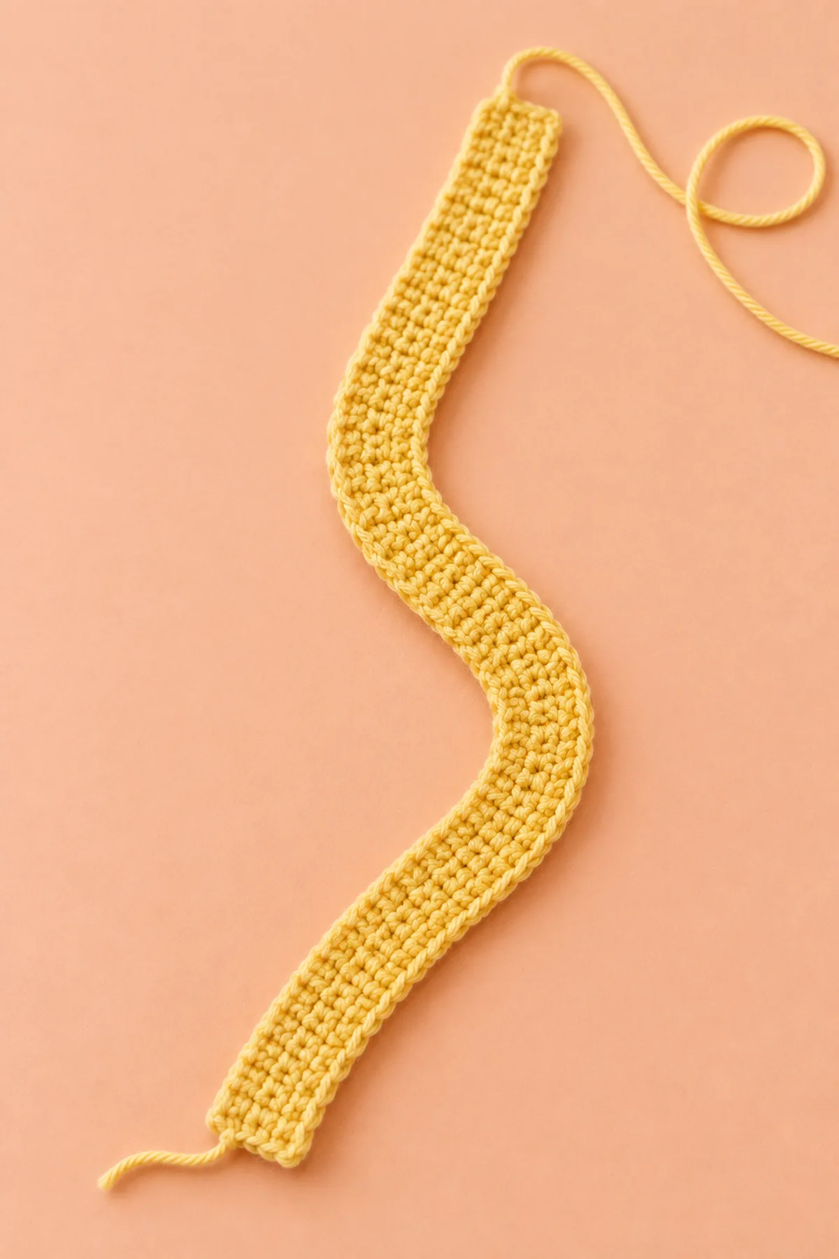 Narrow flat yellow single crochet strip eight stitches wide and sixty rows long loosely curved on peach table, short ends still separate, working yarn nearby; no elastic inserted, no ruffle, no orange or cream