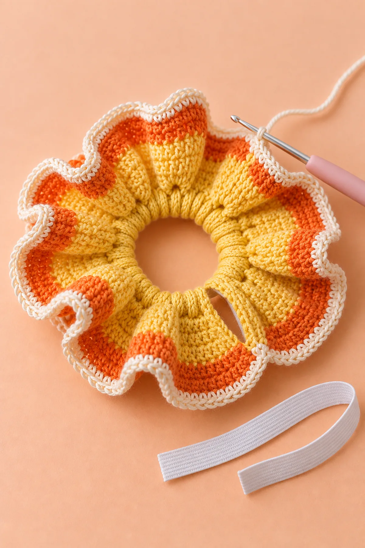 Yellow crochet ring casing with its large yellow-and-orange curly ruffle already worked and narrow cream edging in progress, small hook at cream working edge, service opening on inner casing accessible, separate white flat elastic strip beside it; not worn, no people