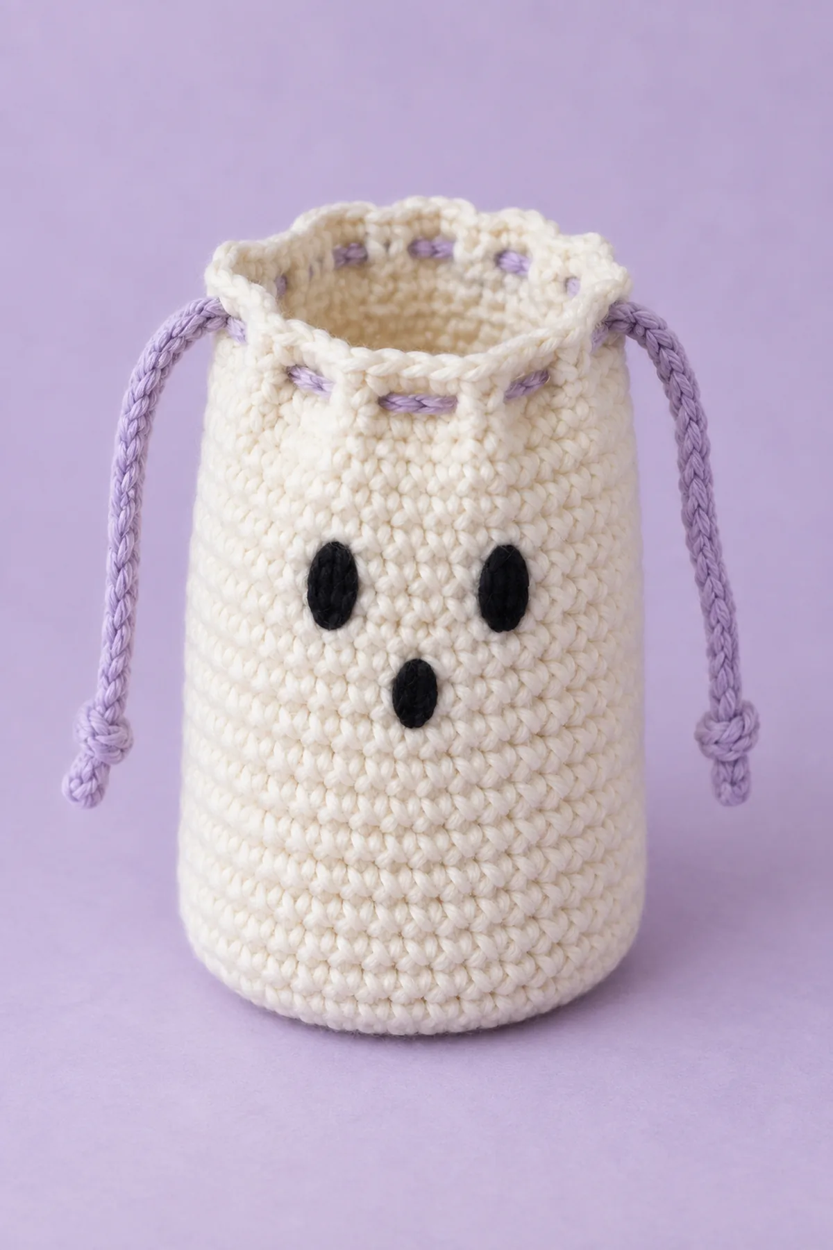 One small cream single-crochet ghost drawstring gift pouch, hollow cylindrical body with flat circular base, two tiny black embroidered oval eyes and smaller oval mouth, two lavender chain drawstrings exiting opposite sides of twelve-eyelet rim. Opening partly open showing empty interior, about 3.4 inches wide and 4.7 inches tall, no stuffing, no arms, no skirt, no beads, pale lavender background