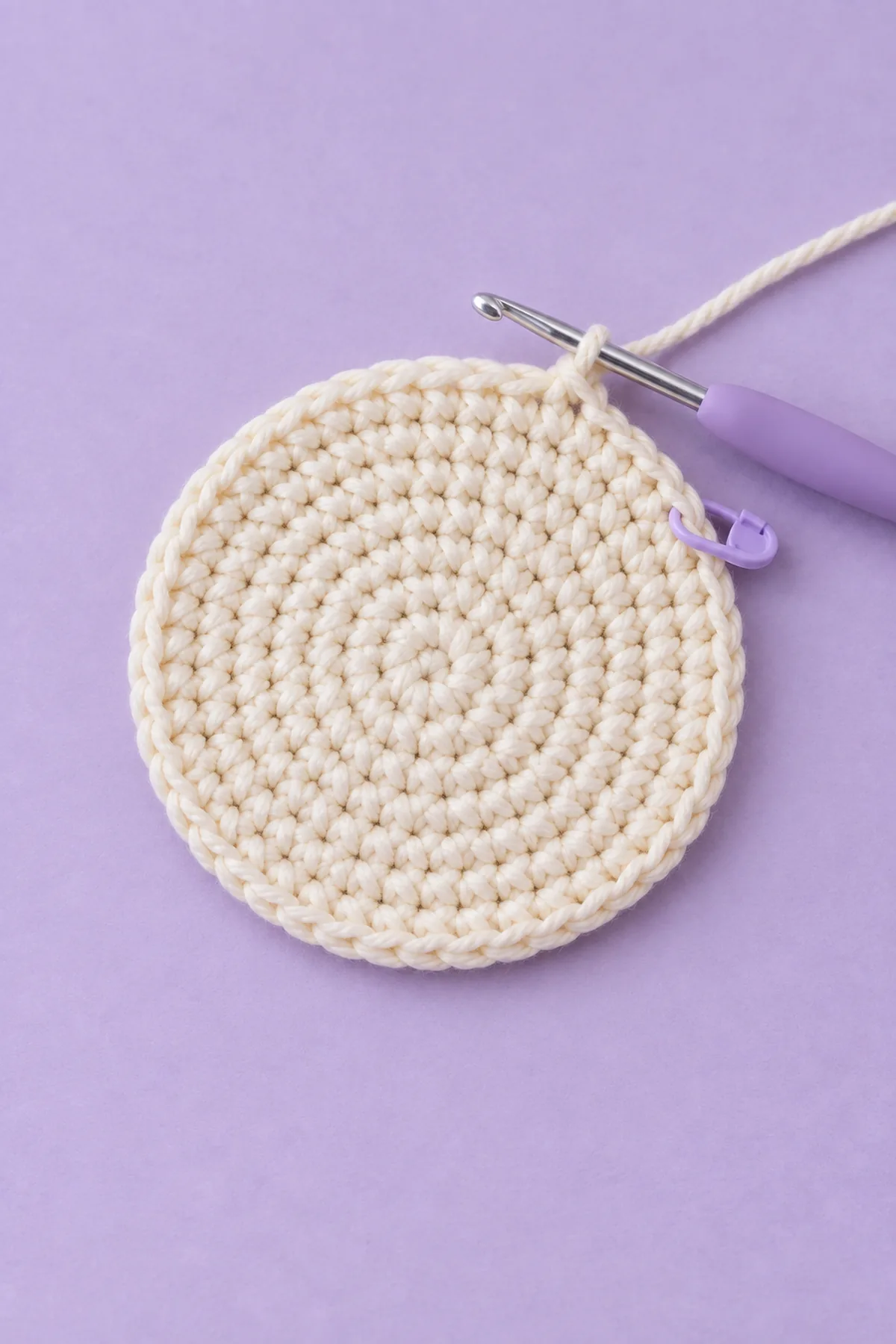 Flat circular cream single-crochet disk after eight increasing rounds, working hook attached at edge and one marker, no walls, no ghost face, no drawstrings, pale lavender surface