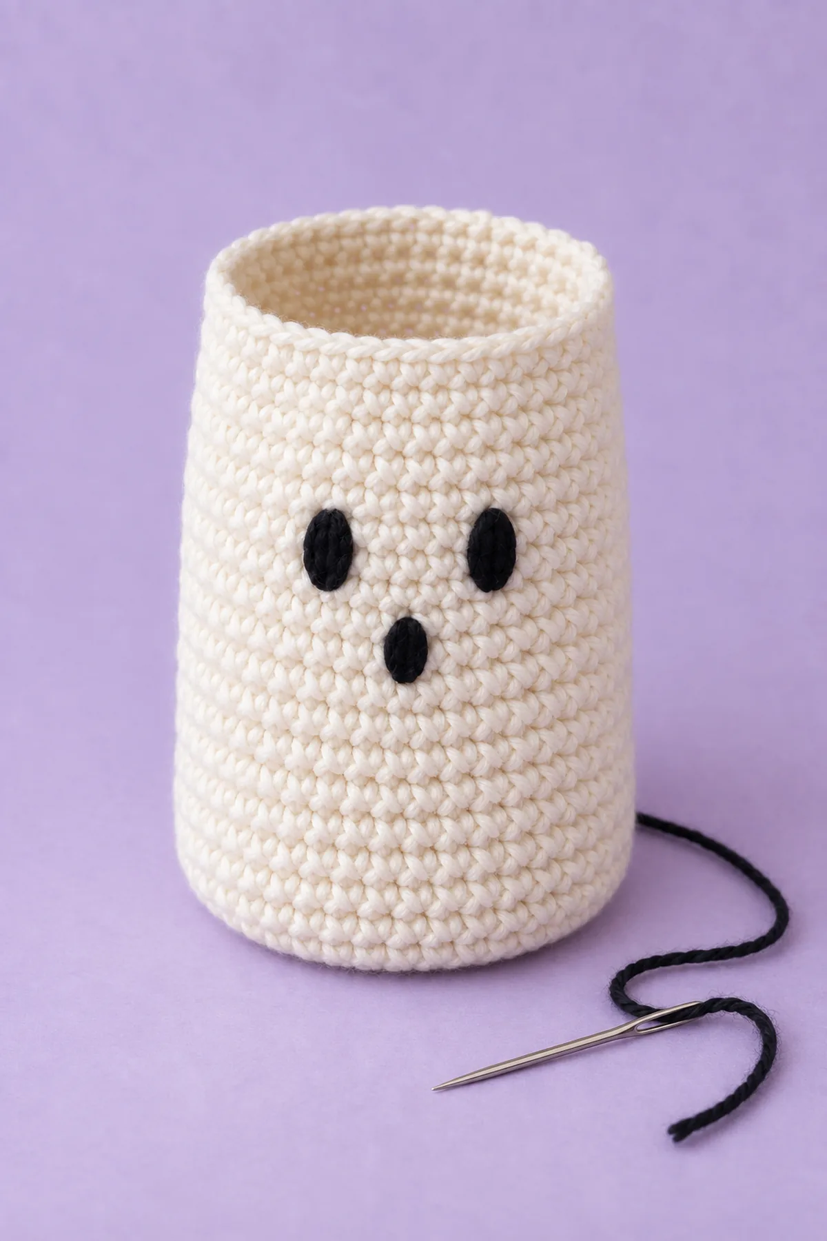 Hollow unfinished cream cylindrical single-crochet pouch with straight plain open upper edge before eyelet rim, black embroidered two eyes and small oval mouth on front, yarn needle and loose black embroidery tail beside it, no lavender cords yet