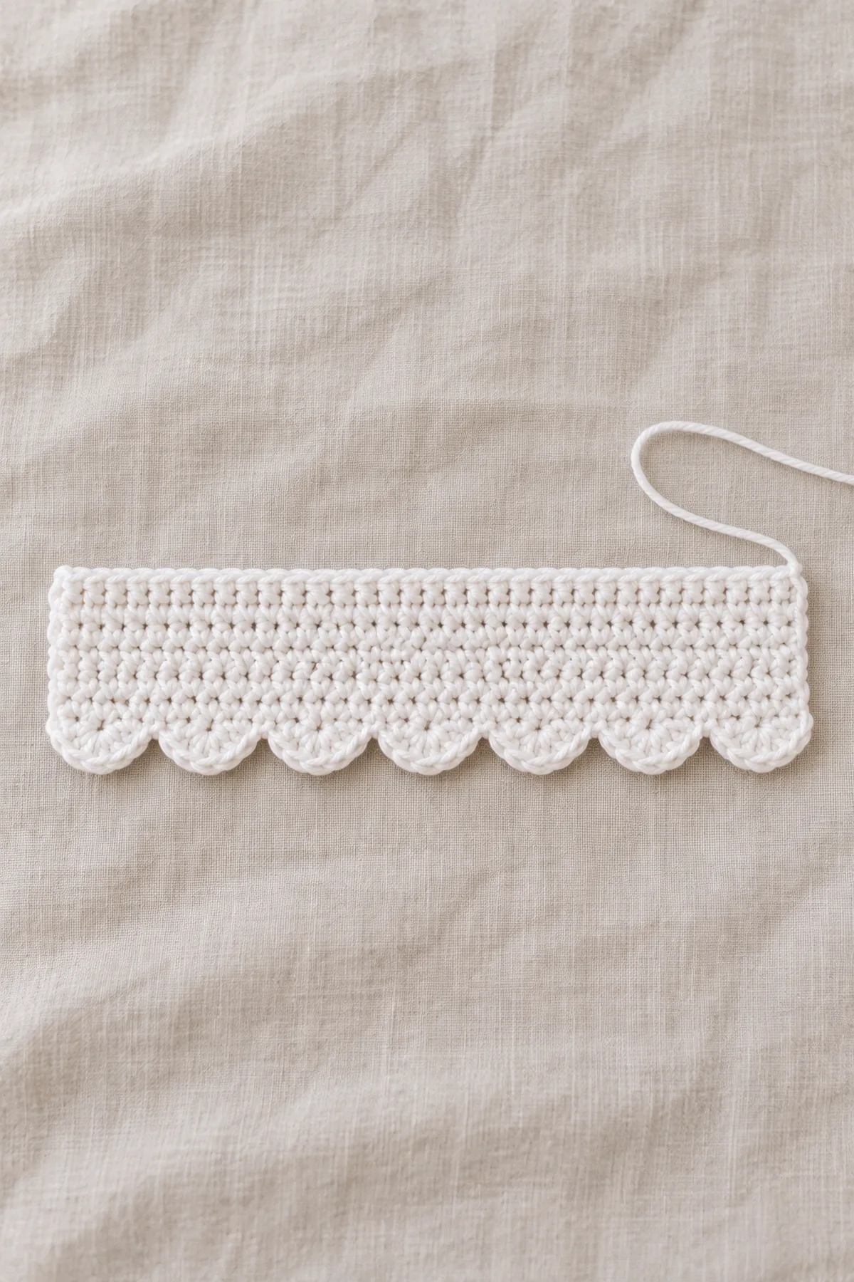 The flat white crochet rectangle now has seven gentle five-double-crochet shell scallops along its lower long edge, straight upper edge, no embroidered face yet, no button yet, laid on pale linen with white yarn tail