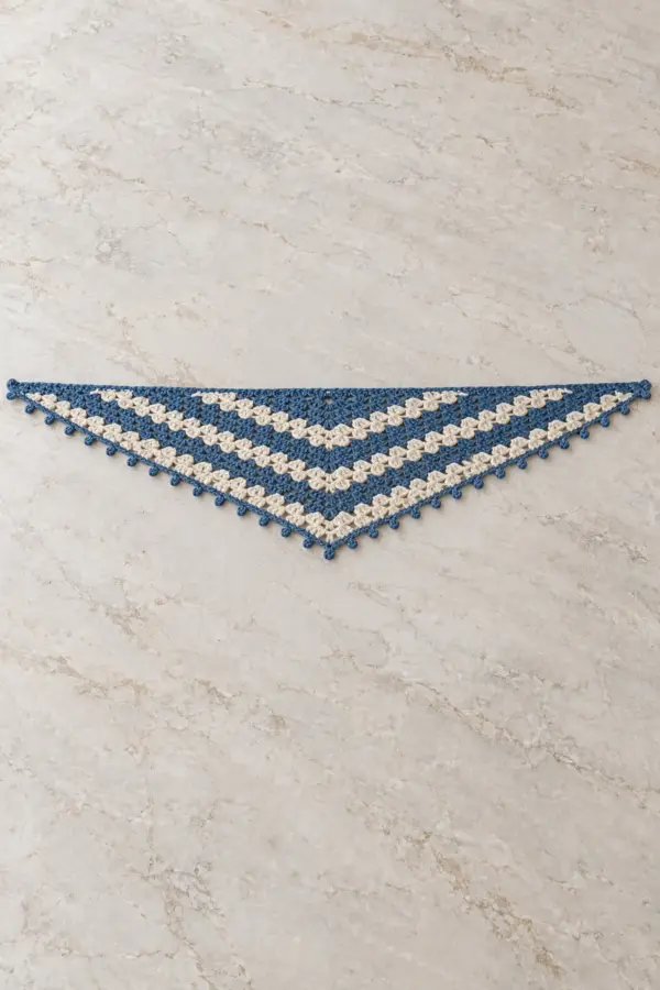 One adult shallow elongated triangular crochet neck scarf laid fully flat on pale stone, wide straight top edge about four times center depth, alternating two-row denim blue and cream granny-cluster stripes forming broad shallow V bands, tiny denim picots only on two angled lower edges, no fringe no hood, no person