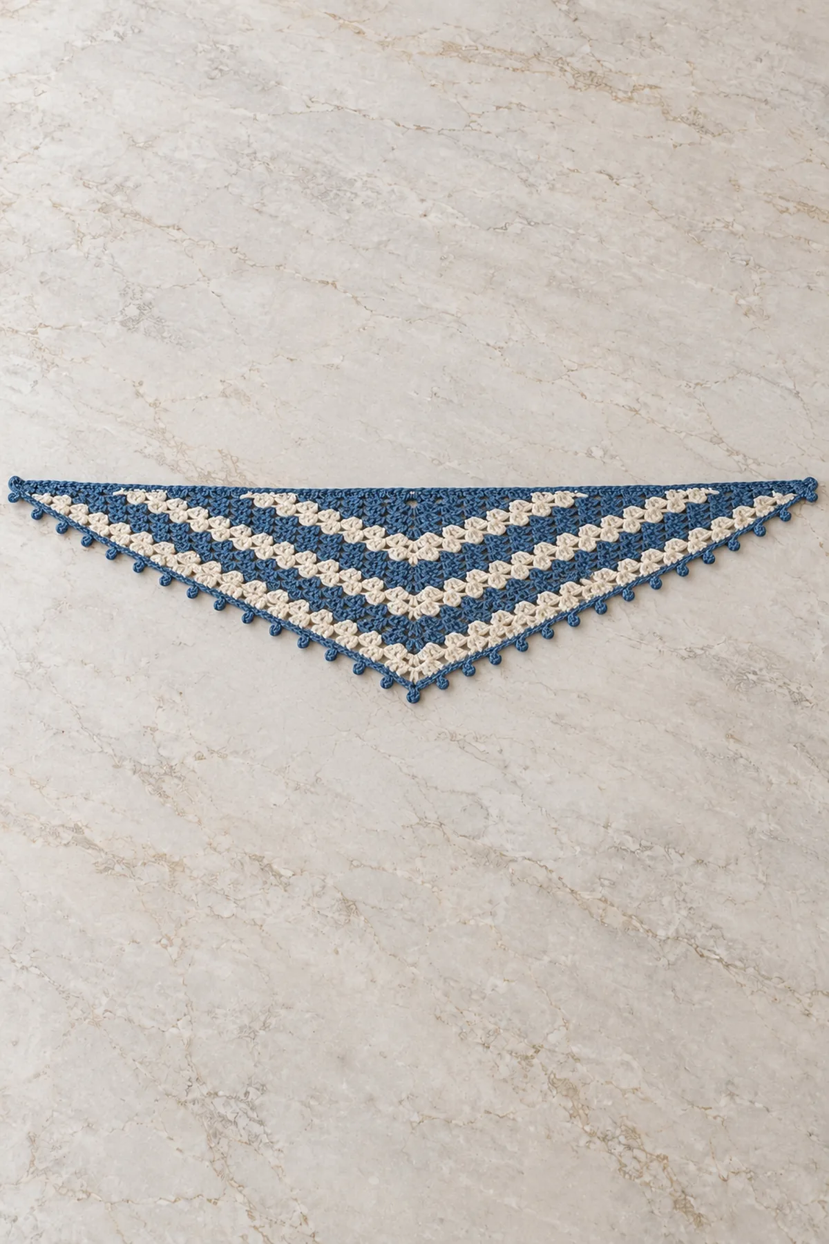 One adult shallow elongated triangular crochet neck scarf laid fully flat on pale stone, wide straight top edge about four times center depth, alternating two-row denim blue and cream granny-cluster stripes forming broad shallow V bands, tiny denim picots only on two angled lower edges, no fringe no hood, no person