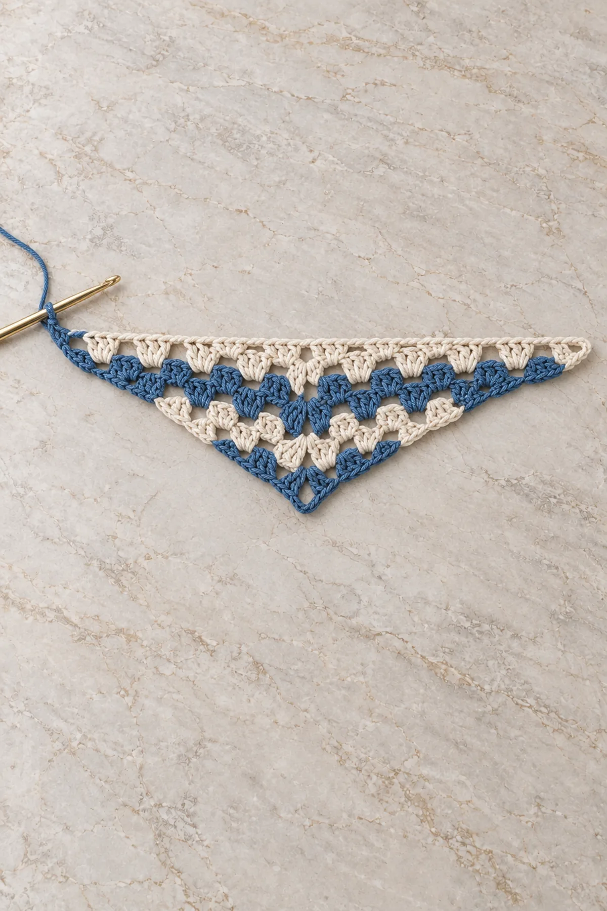 Small unfinished shallow granny crochet triangle with first two denim rows and two cream rows, center chain-space point marked, hook at one wing tip, open groups, no edging yet, pale stone background