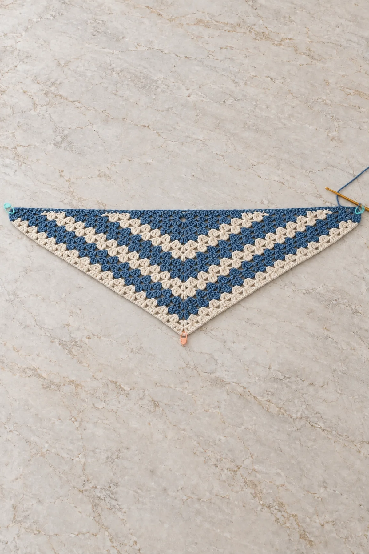 Larger unfinished shallow blue and cream striped granny triangle lying flat, repeated V bands with hook at active outer corner and three markers at tips and center point, no picot edging yet
