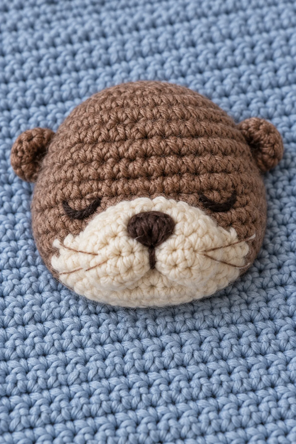 Close-up of the otter's yarn-embroidered closed eyes and ivory muzzle, rounded ear seams, and securely sewn head base on the mist-blue square