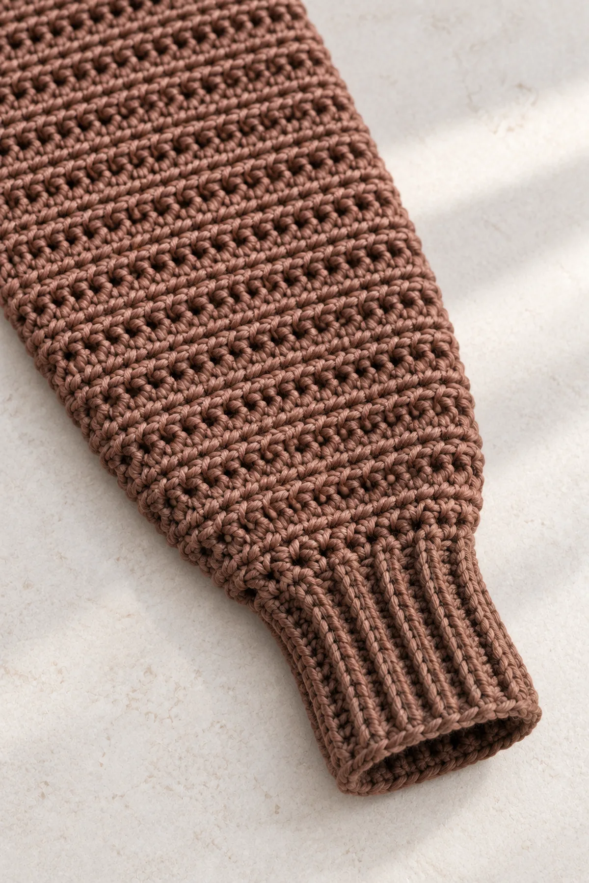 Macro view of one cocoa sleeve end joining a narrower cocoa sc-BLO ribbed cuff, two neat decrease rounds between broader sleeve and cuff, ridged crochet fabric and flexible seam visible, no buttons no fingers