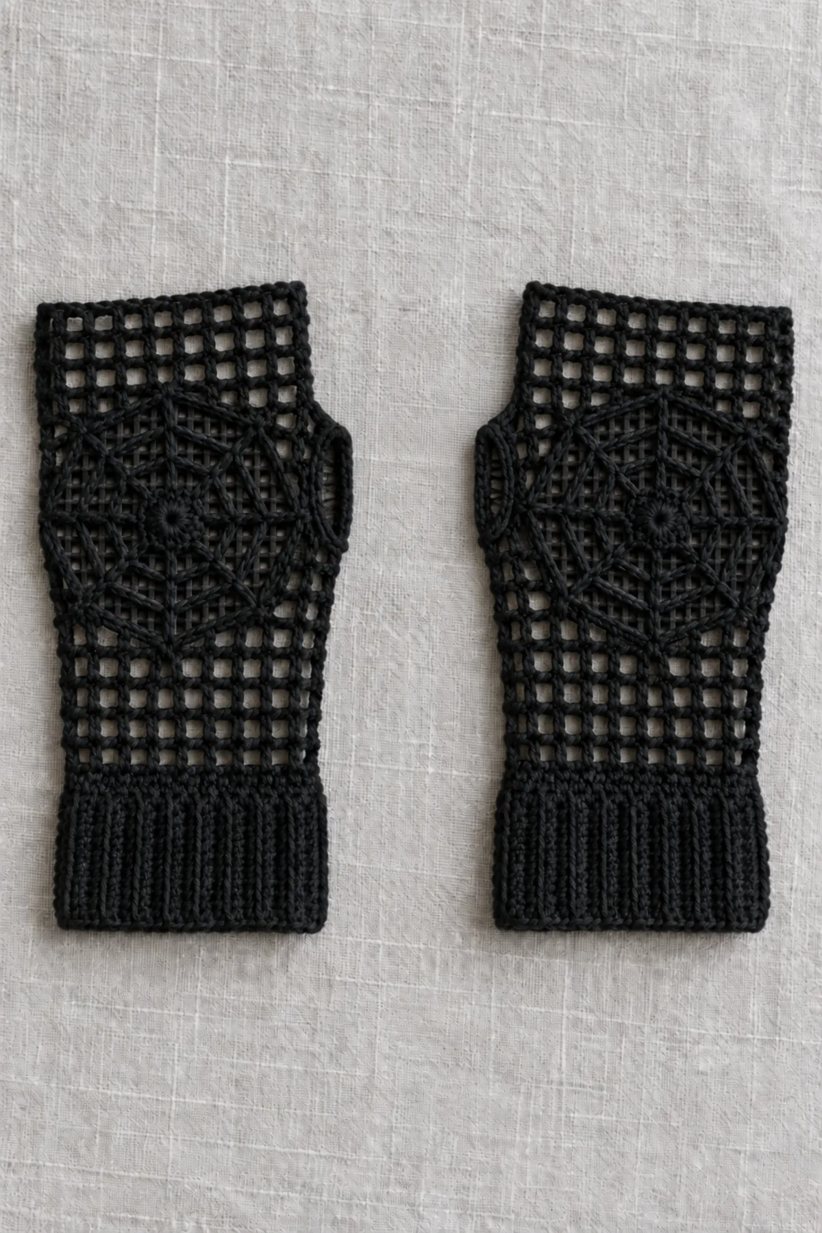 A matched mirrored pair of adult black crochet fingerless gloves, open mesh hands and ribbed cuffs, each with an eight-spoke open spiderweb motif sewn on the back, flat on pale grey linen