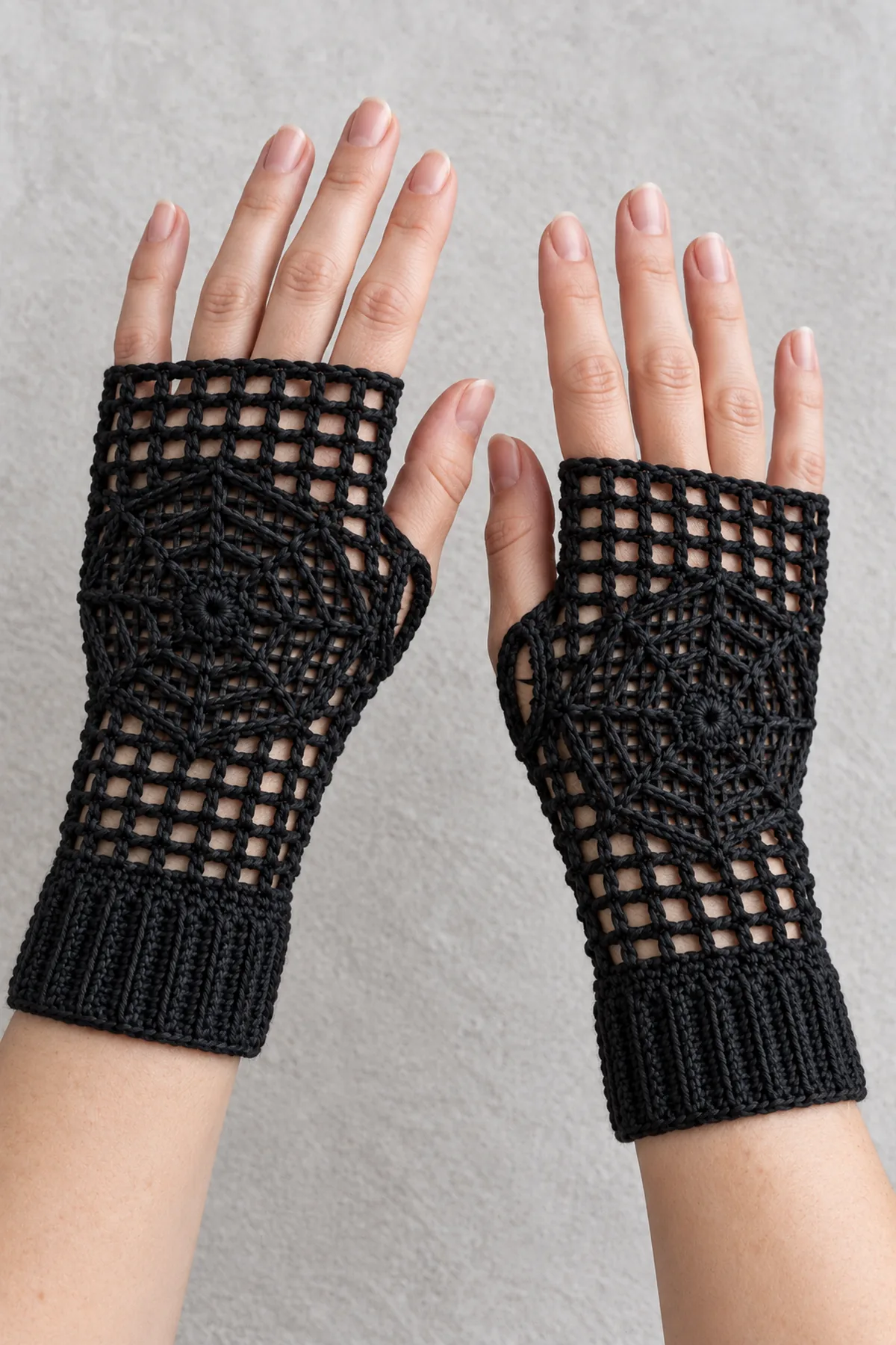 Two adult hands naturally wearing a mirrored pair of black spiderweb fingerless crochet gloves with ribbed cuffs, thumbs through side openings, fingers uncovered, pale grey background
