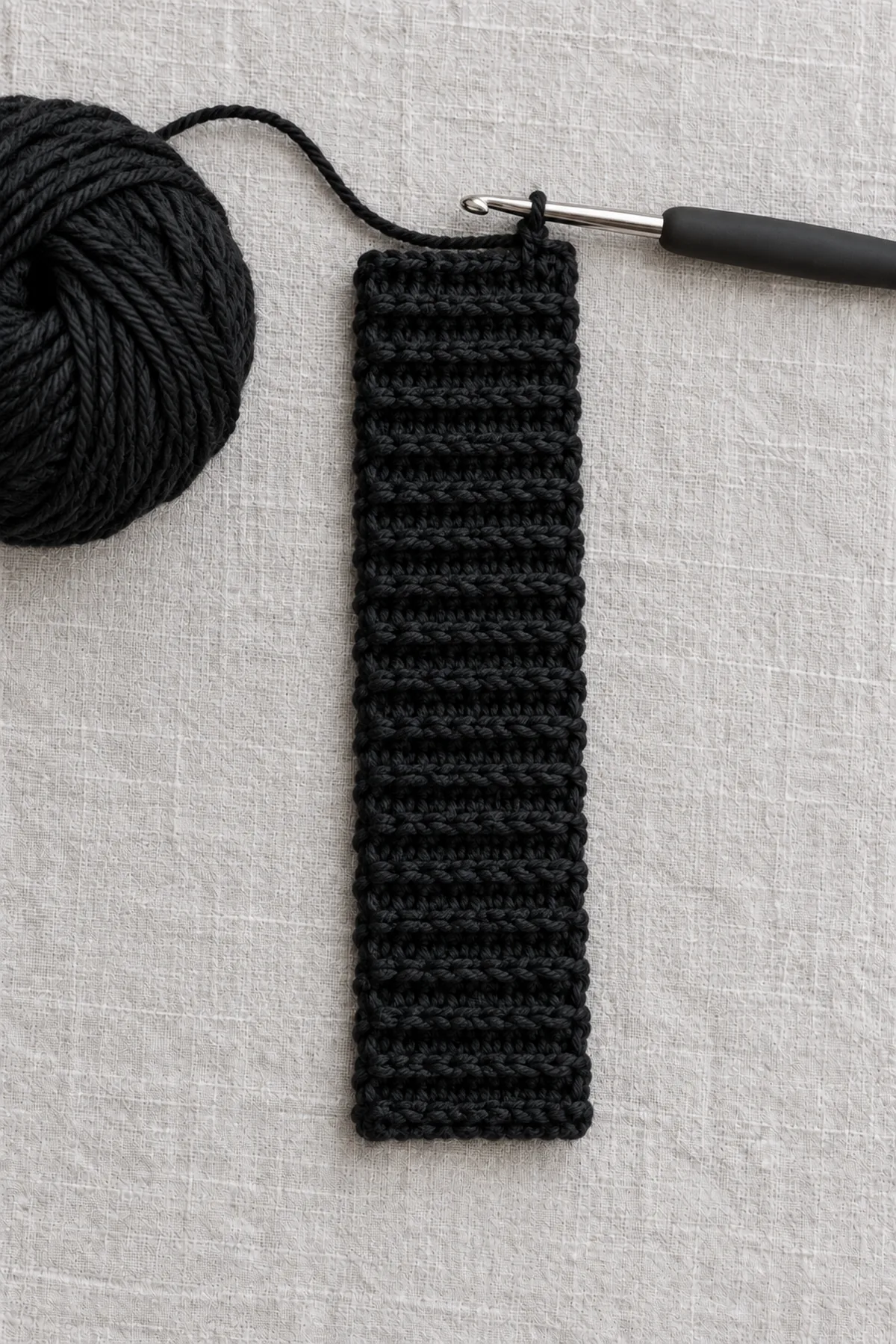 Black eight-stitch-wide back-loop crochet cuff strip with 32 ribbed rows laid flat before joining, hook and markers nearby