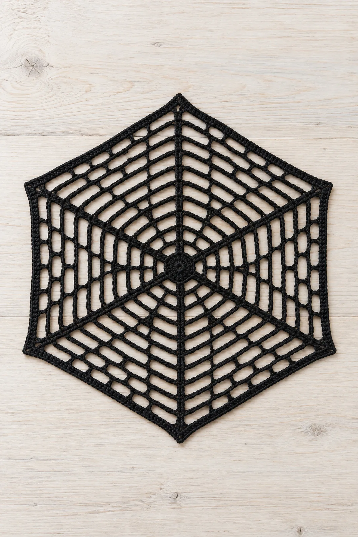 One flat black cotton crochet spiderweb table mat on pale bleached wood, regular HEXAGON with exactly SIX radial rays from small center, twelve open concentric hexagonal chain-arc mesh rounds, fine solid single-crochet perimeter, approximately ten inches point-to-point. Six corners, airy lace, no extra radial spokes, no spider, no plate, no scallops