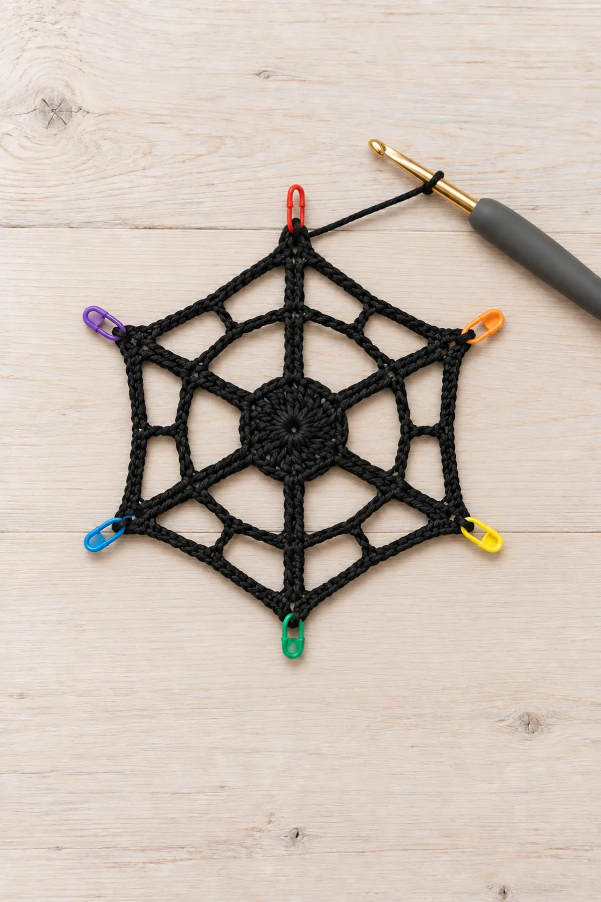 Small unfinished black cotton crochet hexagonal web with exactly SIX rays and ONLY TWO open concentric mesh rounds around a tiny six-stitch center, six colored markers at outer anchor stitches, working hook beside it on pale wood. No full-size mat, no border