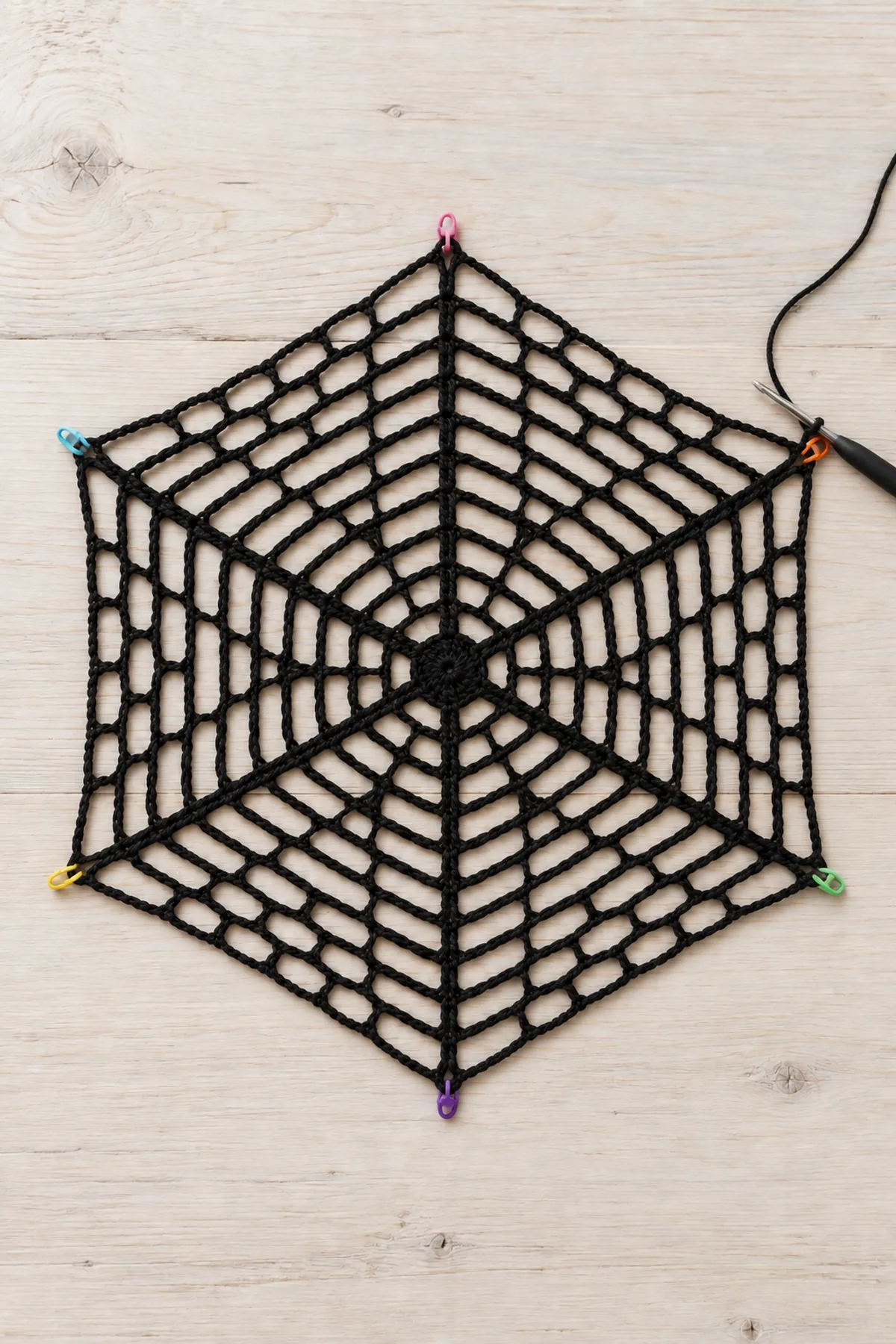 Unfinished flat black cotton crochet hexagonal web after twelve open chain mesh rounds with exactly SIX rays, SIX corners and six colored markers at outer anchors, working black yarn and hook at one corner, no solid outer border yet, pale wood