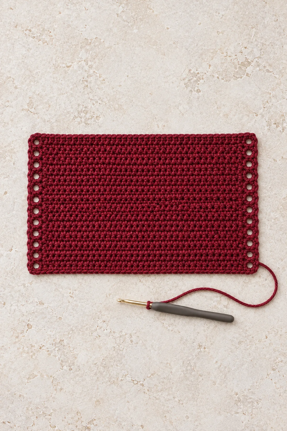 Flat wine-red crochet rectangular panel approximately eight by five inches with fourteen small chain eyelets along each short vertical edge, hook and yarn on pale stone, no bottle inside and no black collar attached