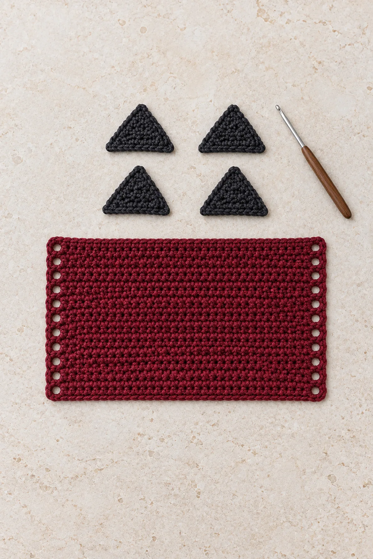 Four separate small identical charcoal crochet triangles arranged as two pairs beside flat wine-red eyelet-edged rectangular panel on pale stone, two triangles partly whipstitched into a double layer, no completed wrap