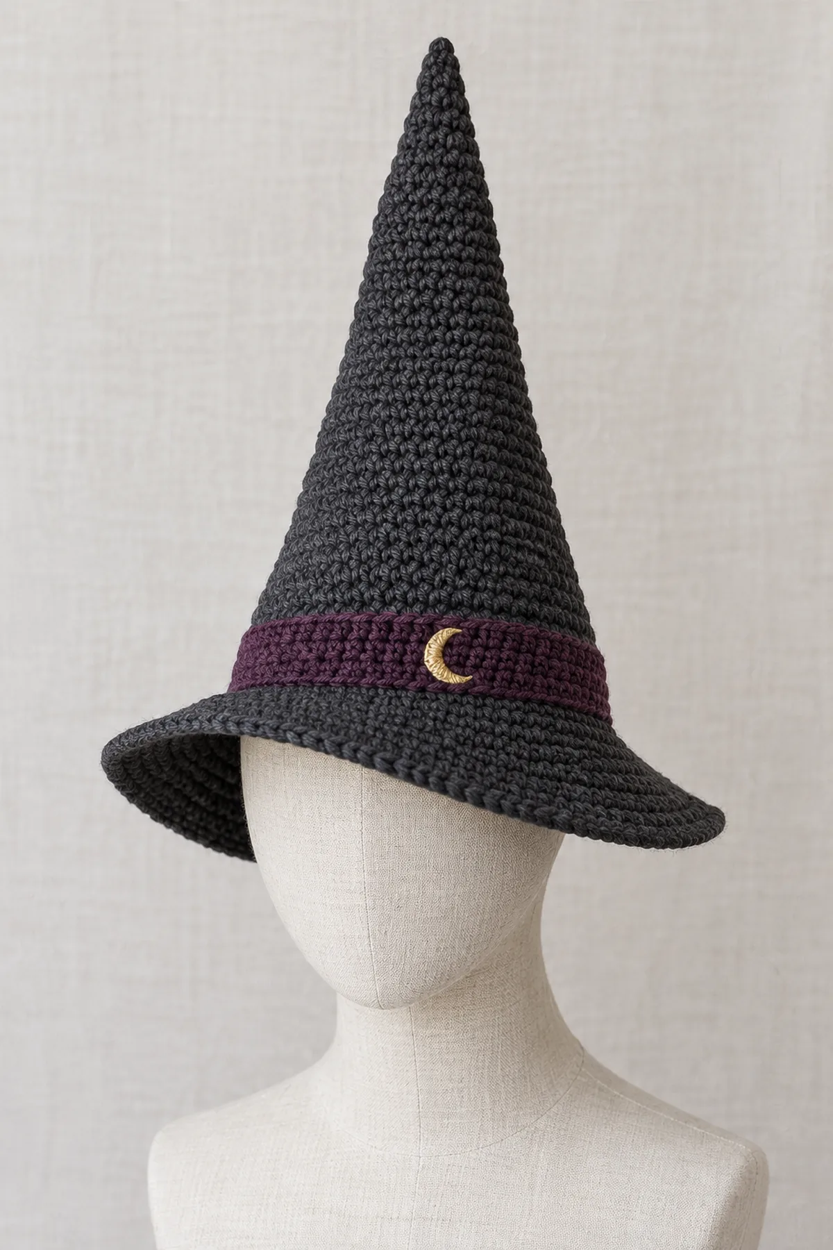 Adult charcoal crochet witch hat with a tall straight tapered cone, plum band with small gold embroidered crescent, and a soft asymmetric brim deeper on one side, on a neutral adult head form