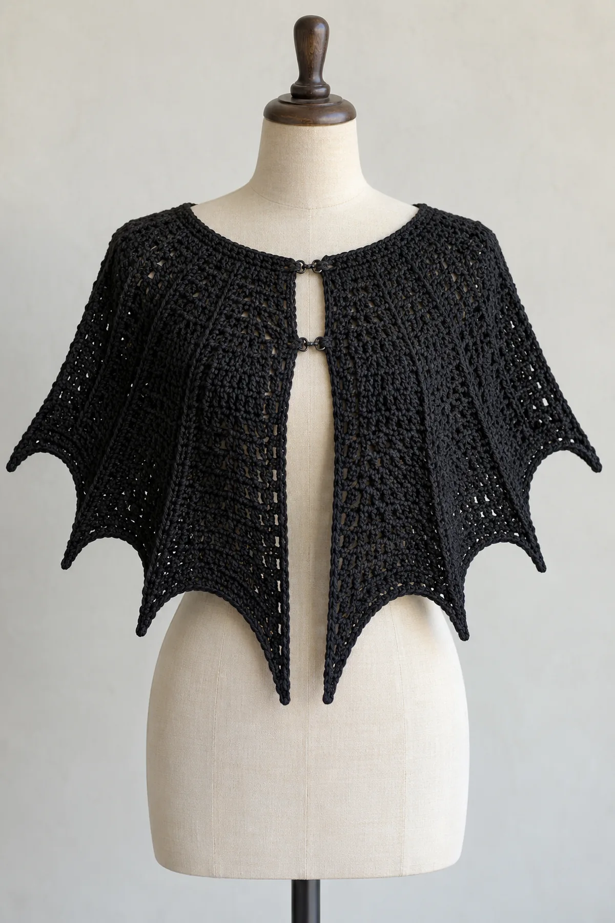 Original adult black crochet batwing shoulder cape on an adult dress form, curved neckline, open front secured with two subtle black hook-and-eye closures, six distinct pointed scalloped wing hem lobes with center pair longest, no hood or sleeves