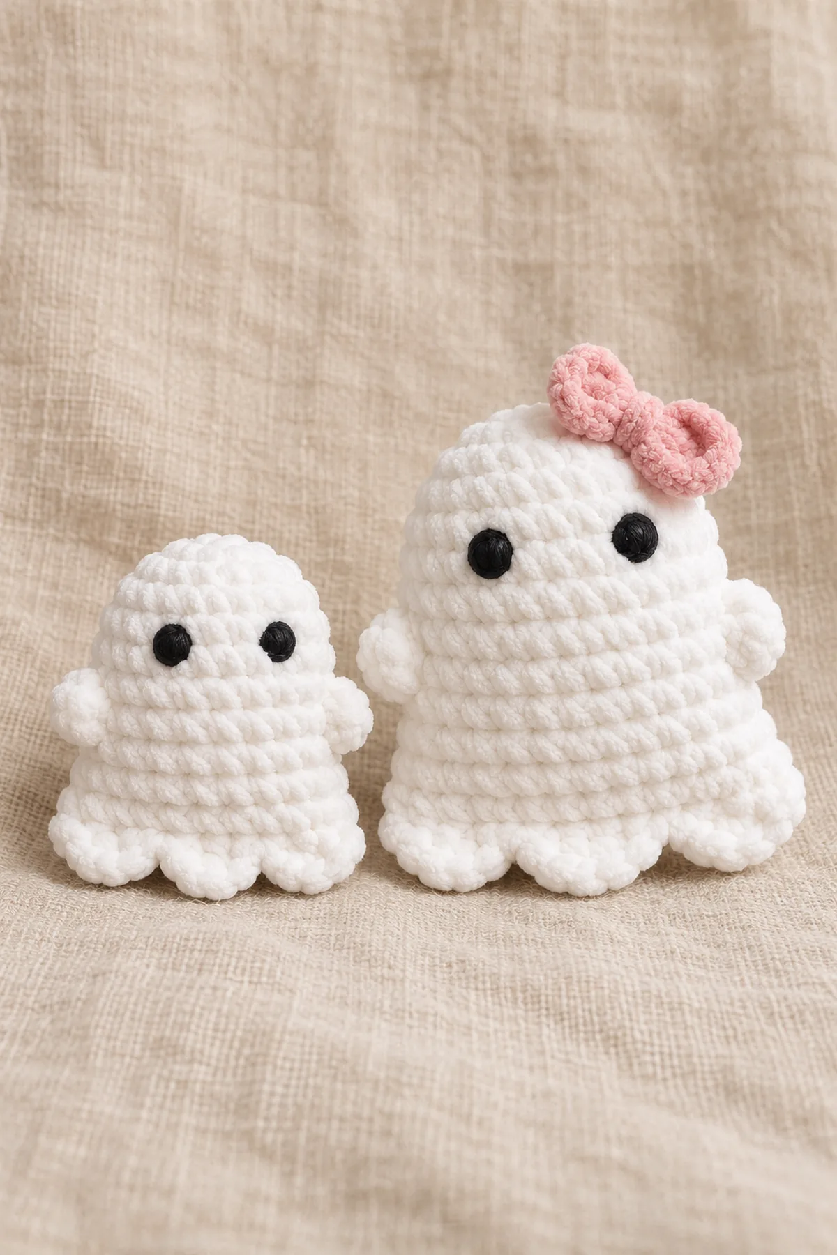 Two white bell-shaped chenille ghosts with a domed top, two stubby arms and exactly three front lower scallops; larger ghost has a fully sewn blush-pink flat bow. Shallow pillow bodies made from two shaped flat panels, not the existing site's spiral pear body or Halloween trio. Two securely embroidered round black eyes, no mouth. Finished object only, original product photograph composition, no source text or logo.