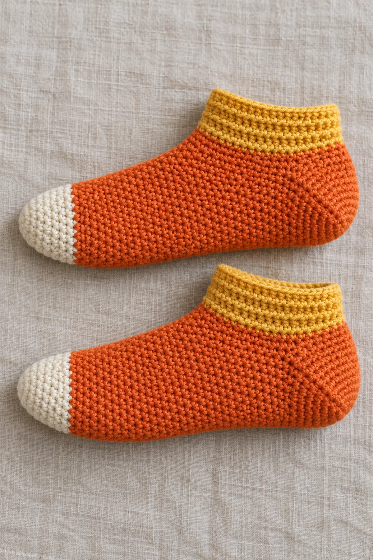 A matching pair of adult ankle-height crochet slipper socks laid flat in side view on oatmeal linen. Cream rounded toes, solid orange feet and clearly cupped orange wedge heels, short yellow cuffs with subtle horizontal back-loop ridges. Fine DK single-crochet texture, fitted foot shape, no face, no laces, no pompom, no baby booties, no people