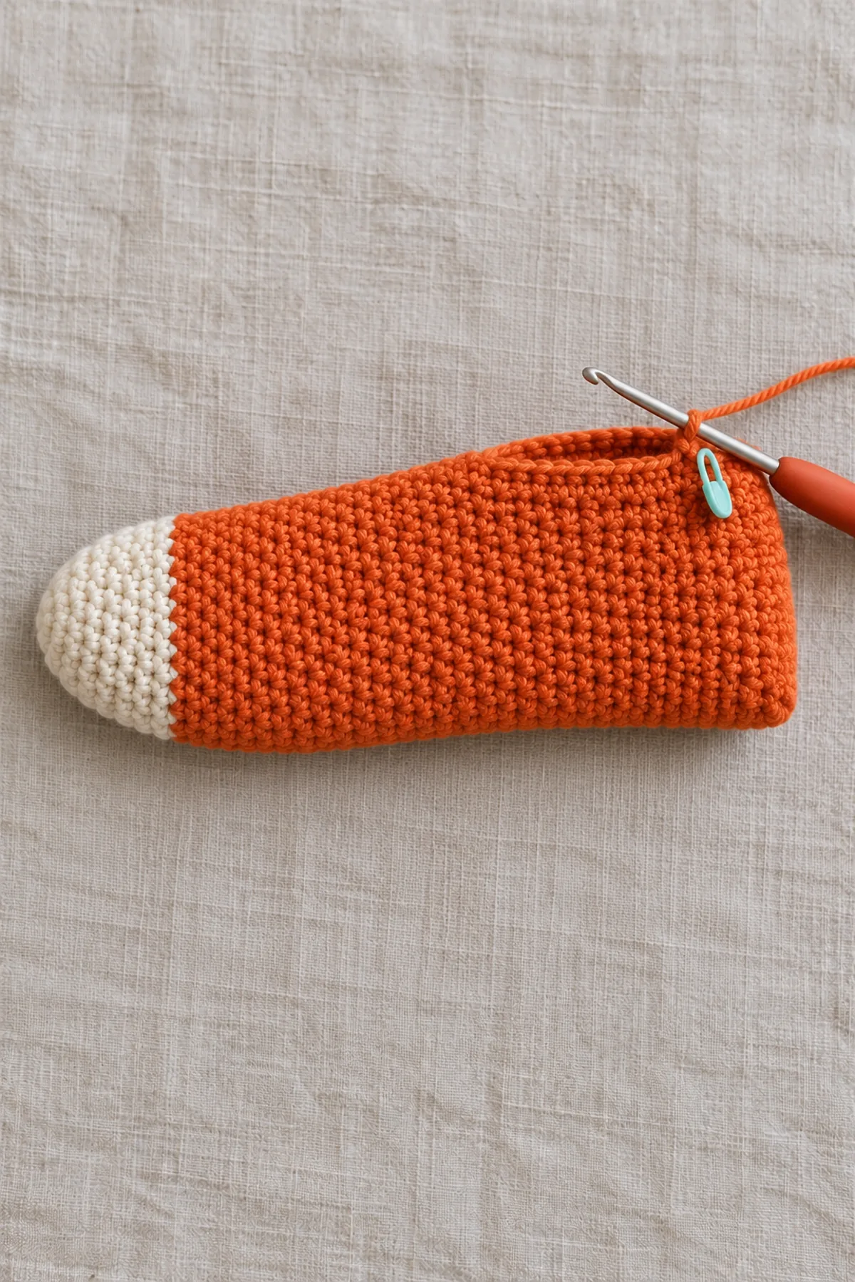 One unfinished adult crochet foot tube: closed cream rounded toe continues into orange straight single-crochet foot, open working edge with hook and marker, no heel opening yet, no yellow cuff, lying flat