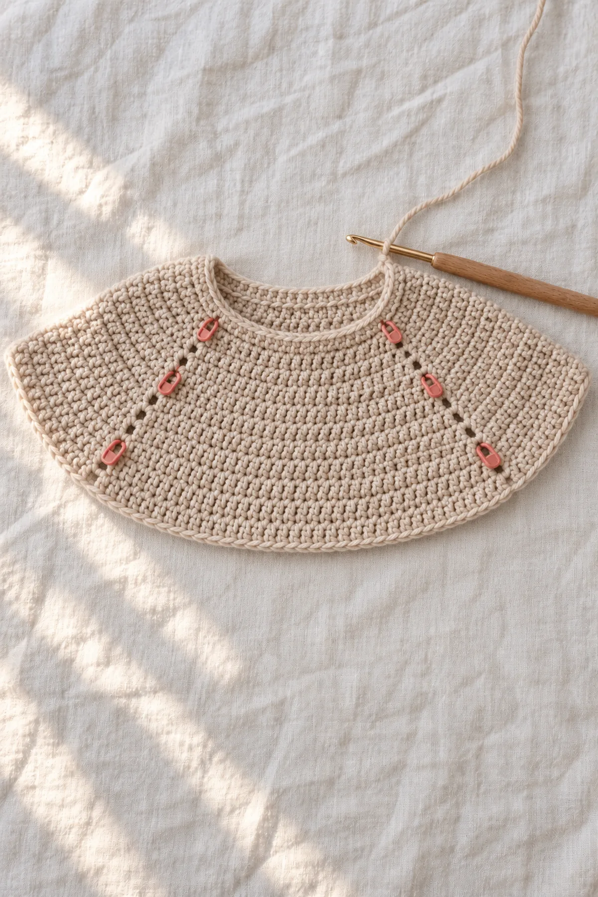 Oat top-down cardigan yoke on hook, flat crescent shape with removable markers showing evenly spaced increase groups; no sleeves or body separation yet.