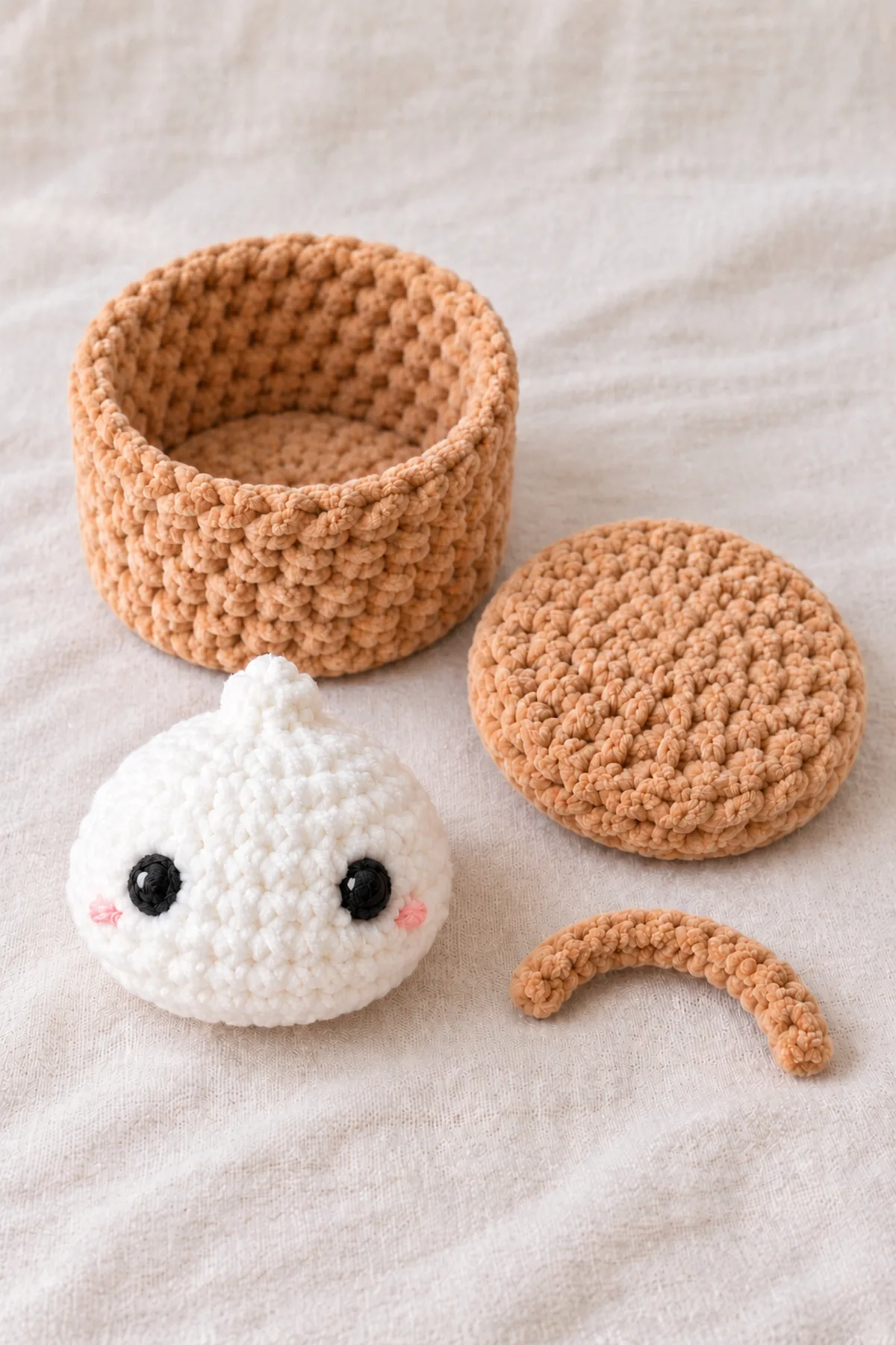 Completed white dumpling with tiny crown, embroidered eyes and blush beside separate open tan cylindrical basket with flat base, separate matching tan shallow overlapping lid and short unsewn tan handle strip.