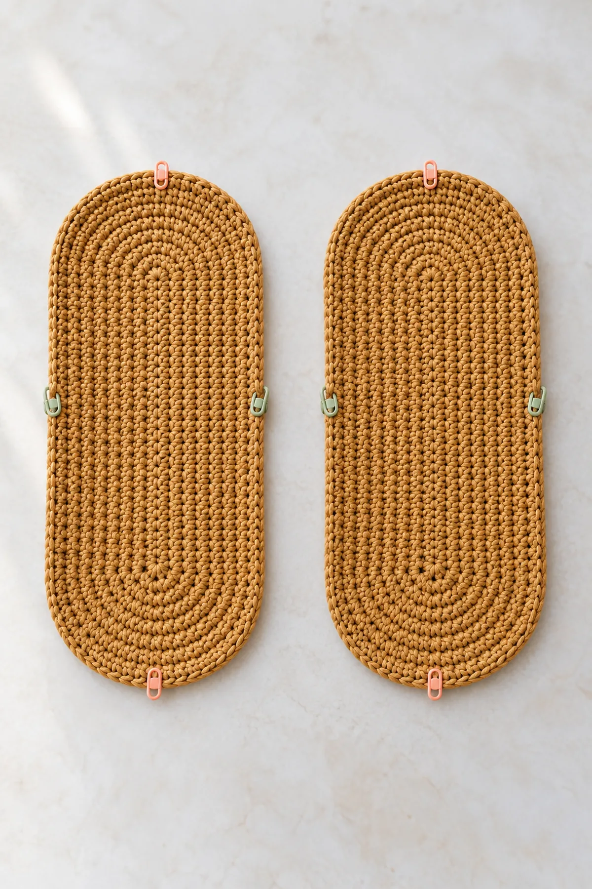 Two separate matching solid warm-ochre crocheted oval bag bases lying flat side by side, stitch markers showing the semicircular ends and long straight sides, no bag walls yet