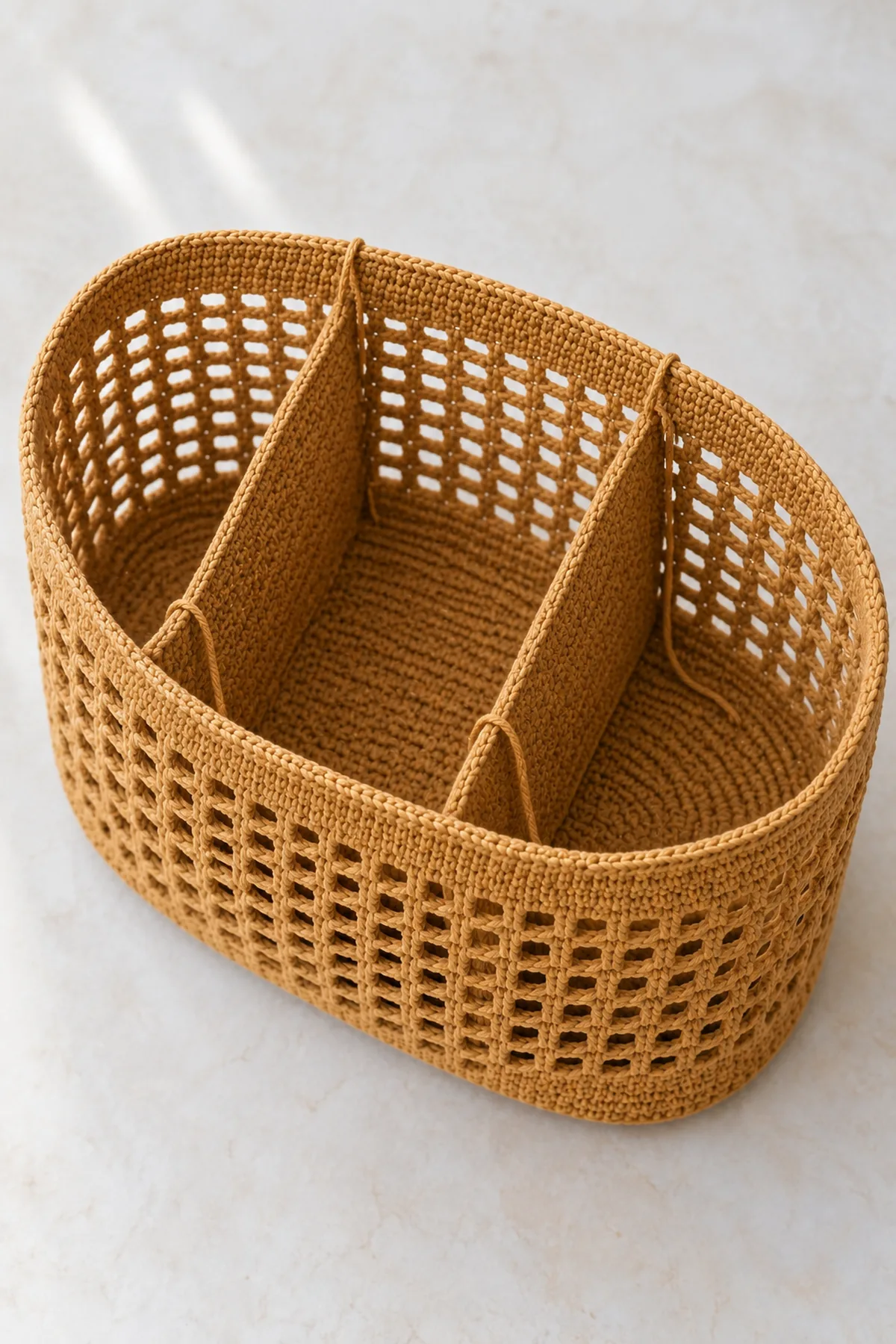 Unfinished warm-ochre cotton mesh market bag with solid oval base and rim but no shoulder handles, open top showing two separate flat crochet divider panels being attached across the interior to form three chambers