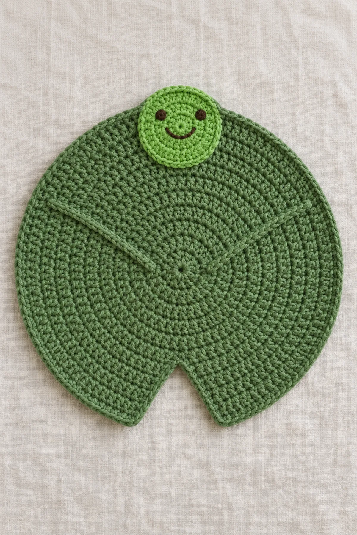 Original leaf-green round crochet lily-pad lovey spread flat, one unmistakable shallow V-shaped wedge notch at one edge, two stitched radial leaf ribs, one small flat bright-green embroidered frog face attached at the opposite edge. No limbs, stuffing or plastic eyes.