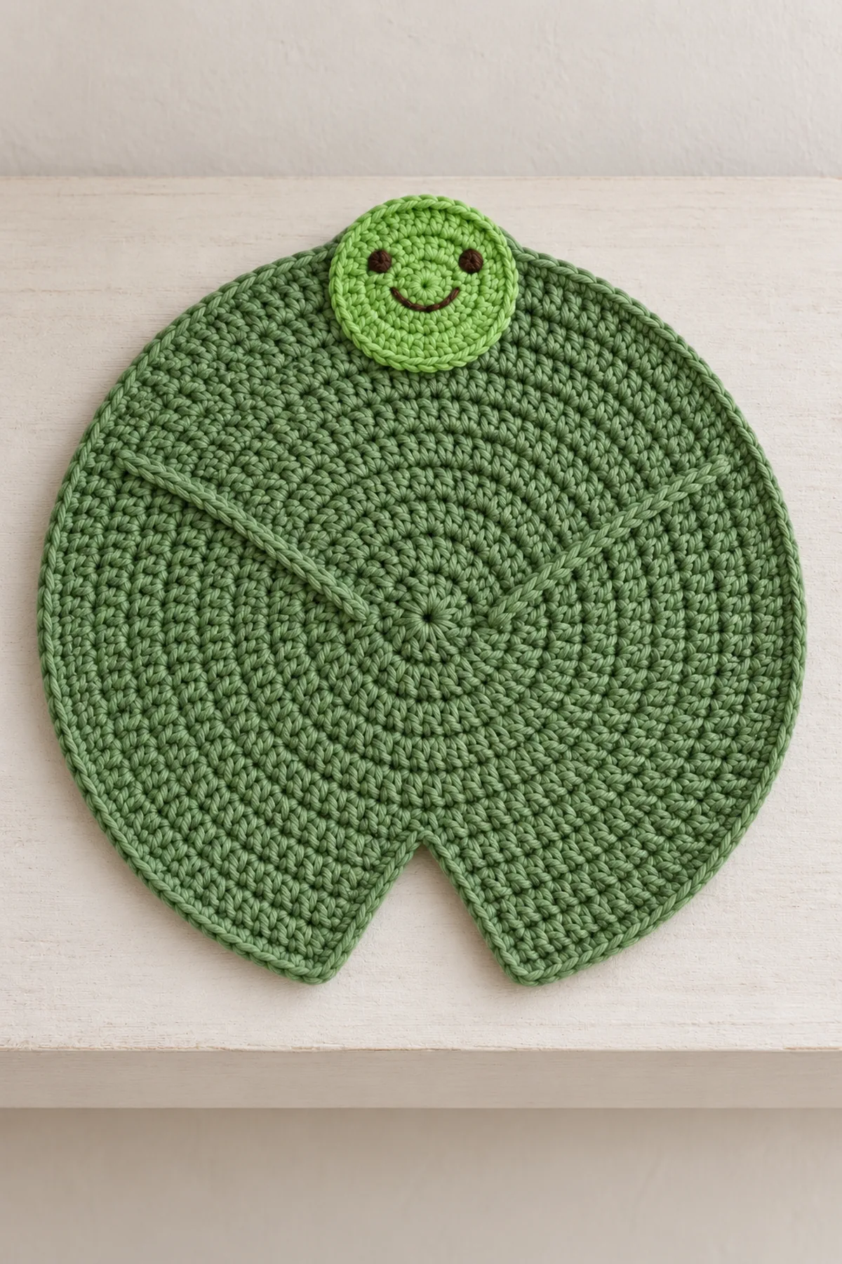 Same green lily-pad crochet lovey displayed flat on a neutral shelf, open notch and opposite flat embroidered frog face fully visible, no baby or crib.