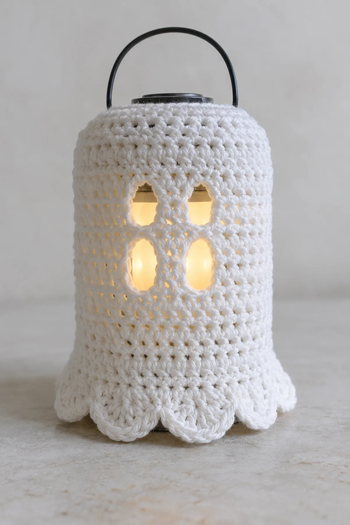 Original white cotton crochet ghost sleeve fitted around a small enclosed cool LED lantern, simple cylindrical shape, two pairs of openwork eye gaps aligned vertically and eight soft scallops at bottom, unlit neutral backdrop, no mouth or glued parts