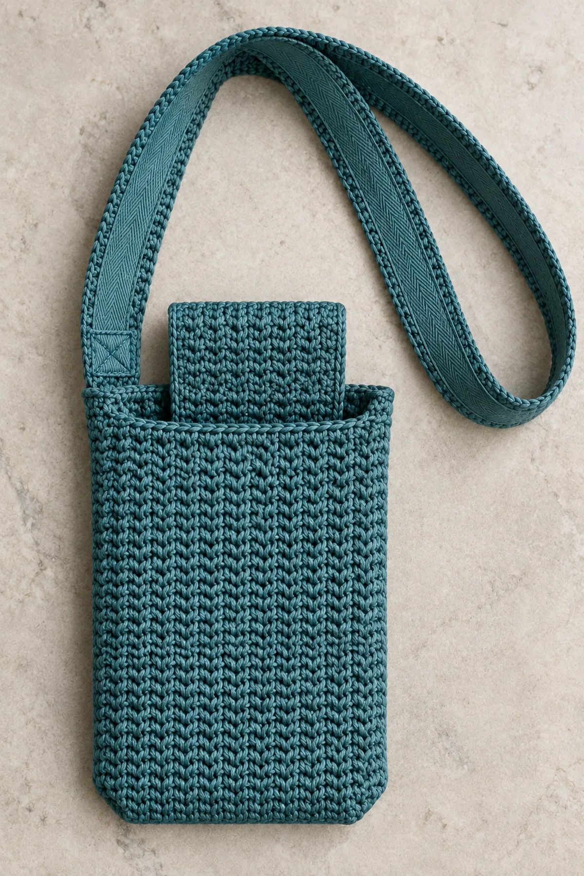 Close view of teal wide flat crochet phone sling strap underside backed by flat cotton twill tape, neat sewing along both tape edges, one strap end attached across upper side seam with rectangle-and-X stitching. Opening and plain tuck flap visible, no hardware