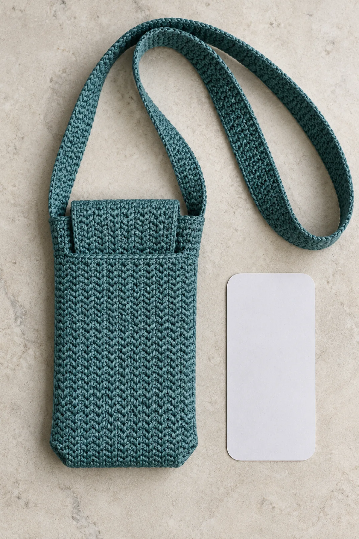 Adult teal fine cotton herringbone crochet phone sling, slim rectangular pocket about 4 by 7.5 inches, two subtly boxed bottom corners and plain integral rectangular tuck flap partly tucked into front opening. Long broad flat 1.6-inch teal strap attached at two upper side seams, arranged as large loop beside pouch. No hardware, fringe, button, ornaments or people. Pale stone background, phone-sized plain paper template beside it
