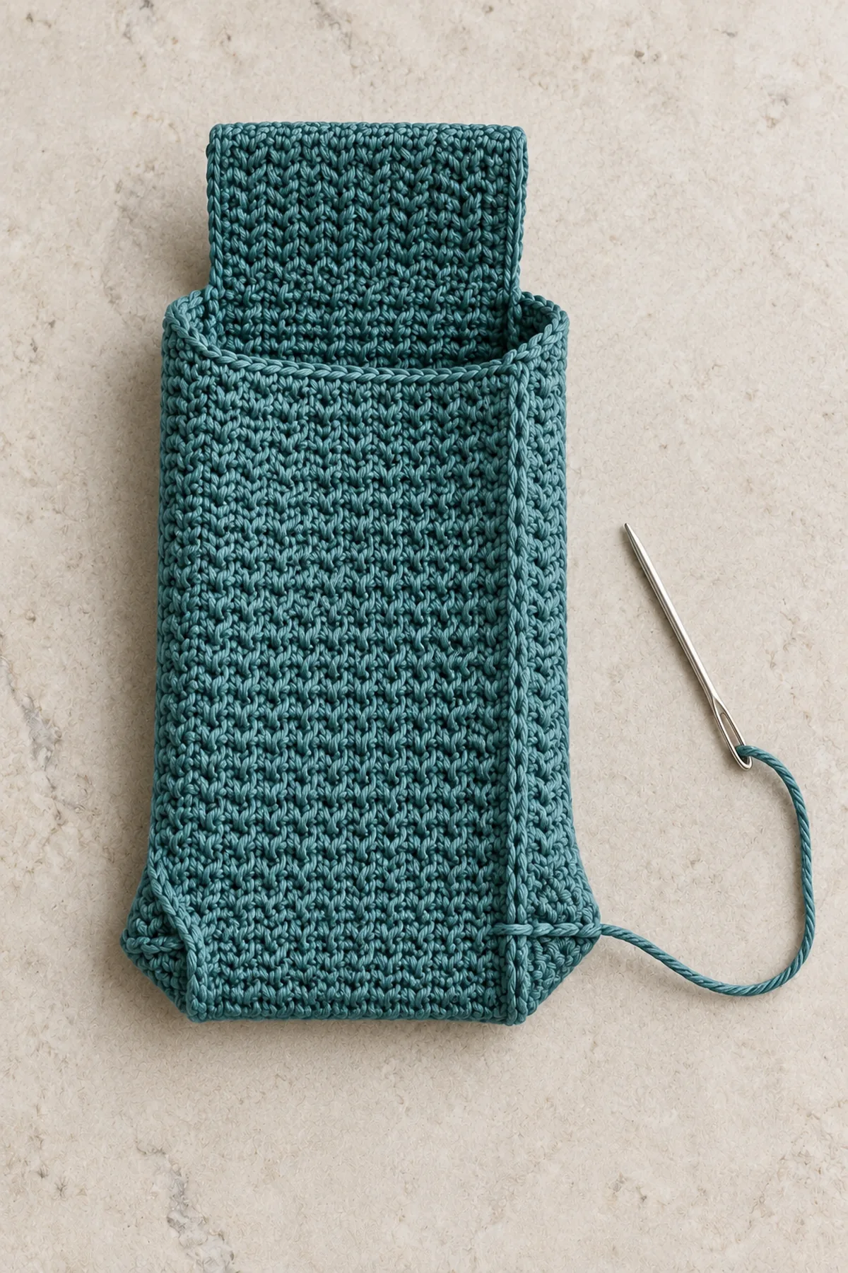 Inside-out small teal crochet phone pouch on pale table, bottom corner flattened with side seam aligned over bottom seam and a short half-inch transverse sewing line forming a small triangular corner tab. Other lower corner already boxed. Integral top flap open, no strap attached yet, blunt needle beside corner