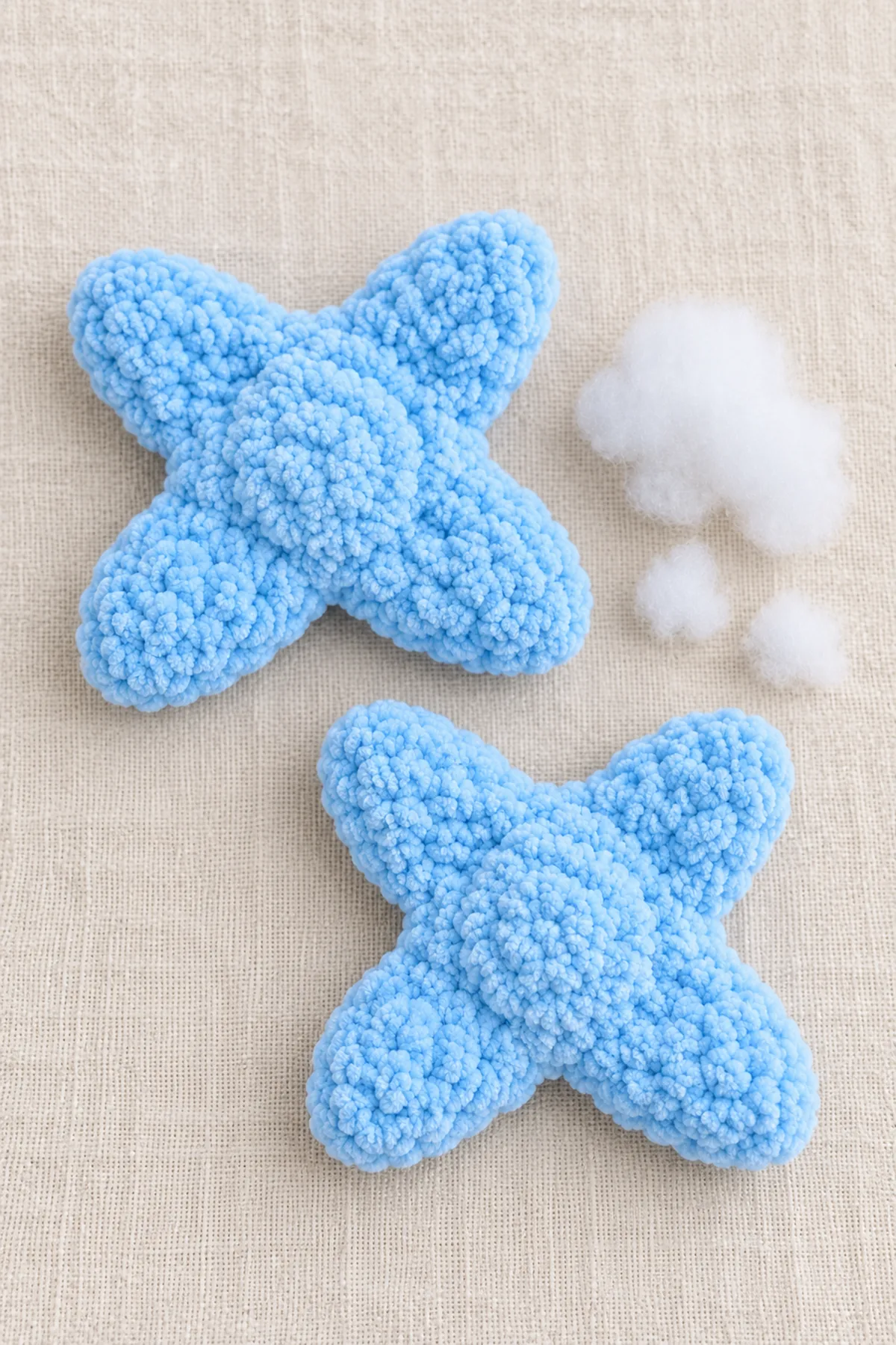 Two powder-blue chenille four-point X-shaped flat star body panels laid separate, one with a few small stuffing tufts beside it; no head, hood, face or assembled doll yet.