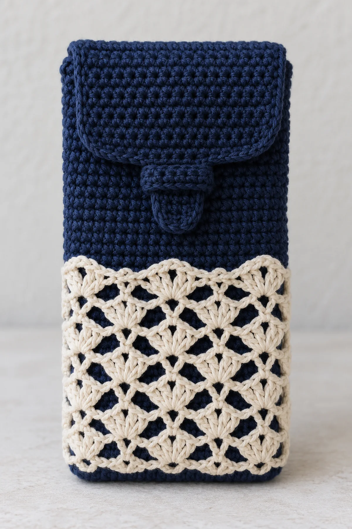 Original navy cotton crochet close-fitting smartphone sleeve standing upright, separate cream openwork lace overlay on lower front, dense navy fold-down flap with narrow integral tab tucked beneath a small front keeper tunnel, no buttons, strap or hardware