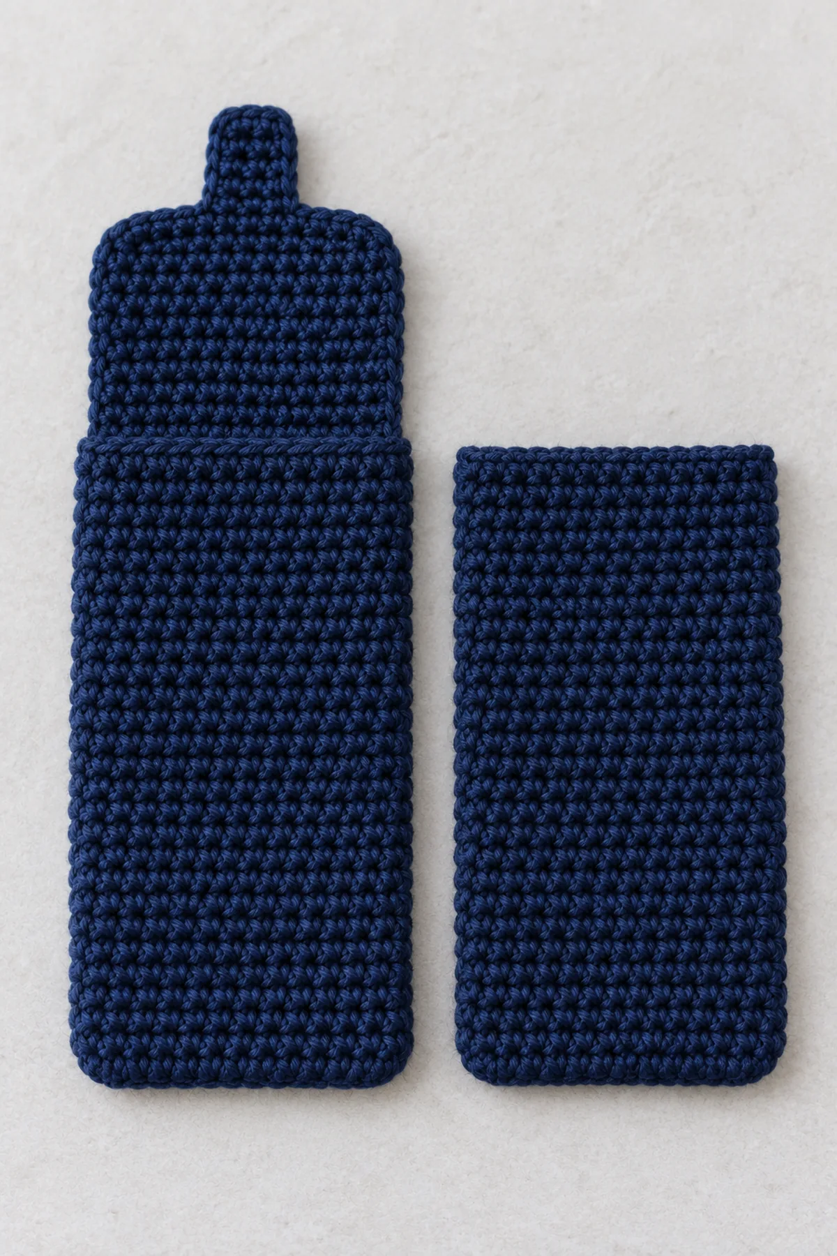 Two separate flat dense navy single-crochet phone sleeve panels, one taller with a fold-down extension and narrow integral tab, no seams or lace yet