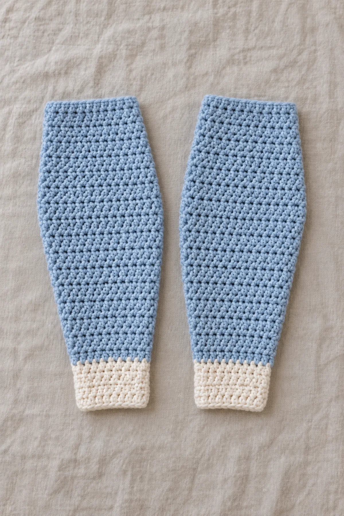 Two separate long blue crochet sleeves taper gently from wide straight upper edges to narrower pale ecru cuffs, lying beside each other unsewn; no body pieces or finished garment.