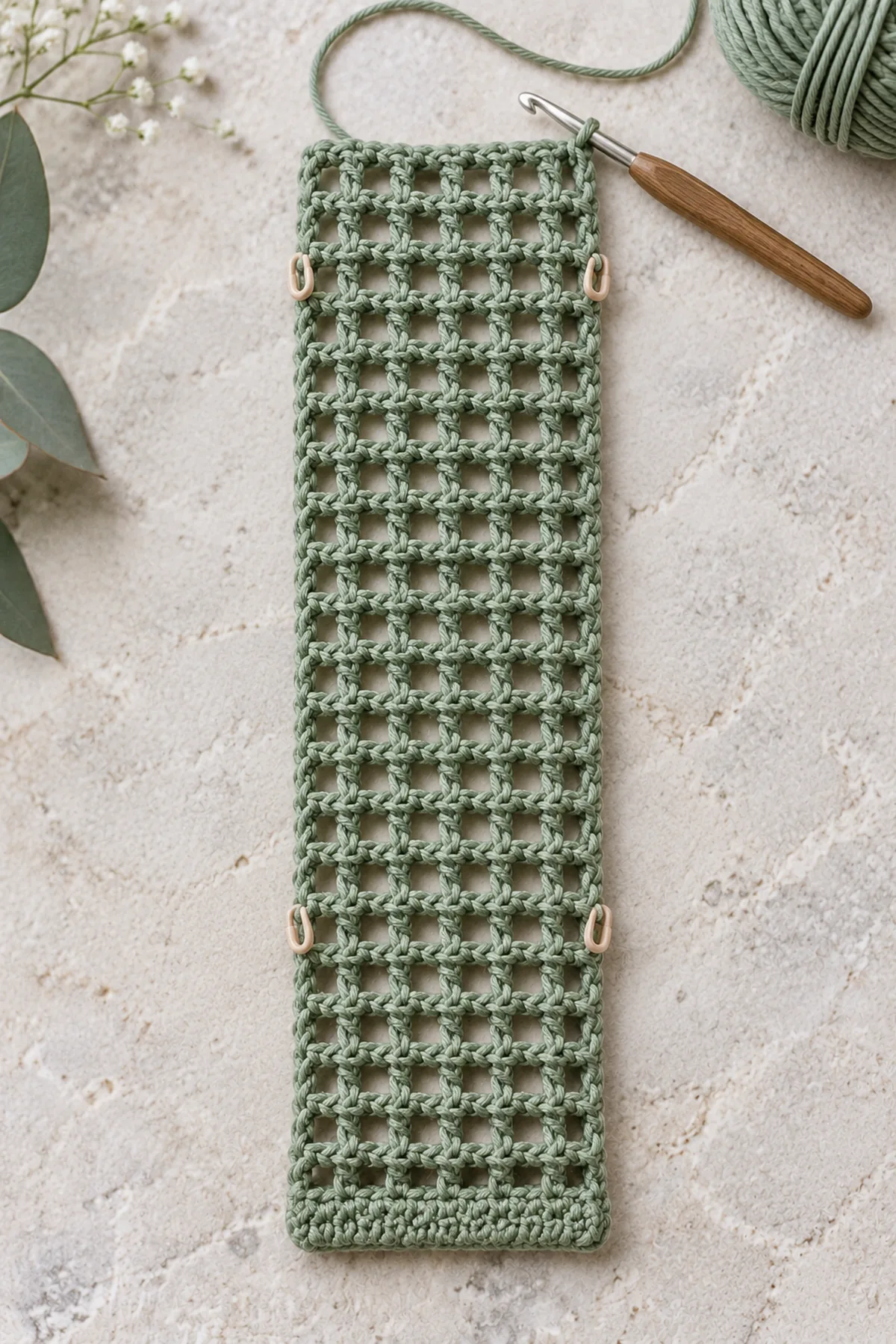 Long narrow unfinished sage crochet mesh rectangle with solid starting rim, nineteen horizontal positions and fold markers at side edges, live working yarn and hook, strip still unfolded and unseamed, no drawstring