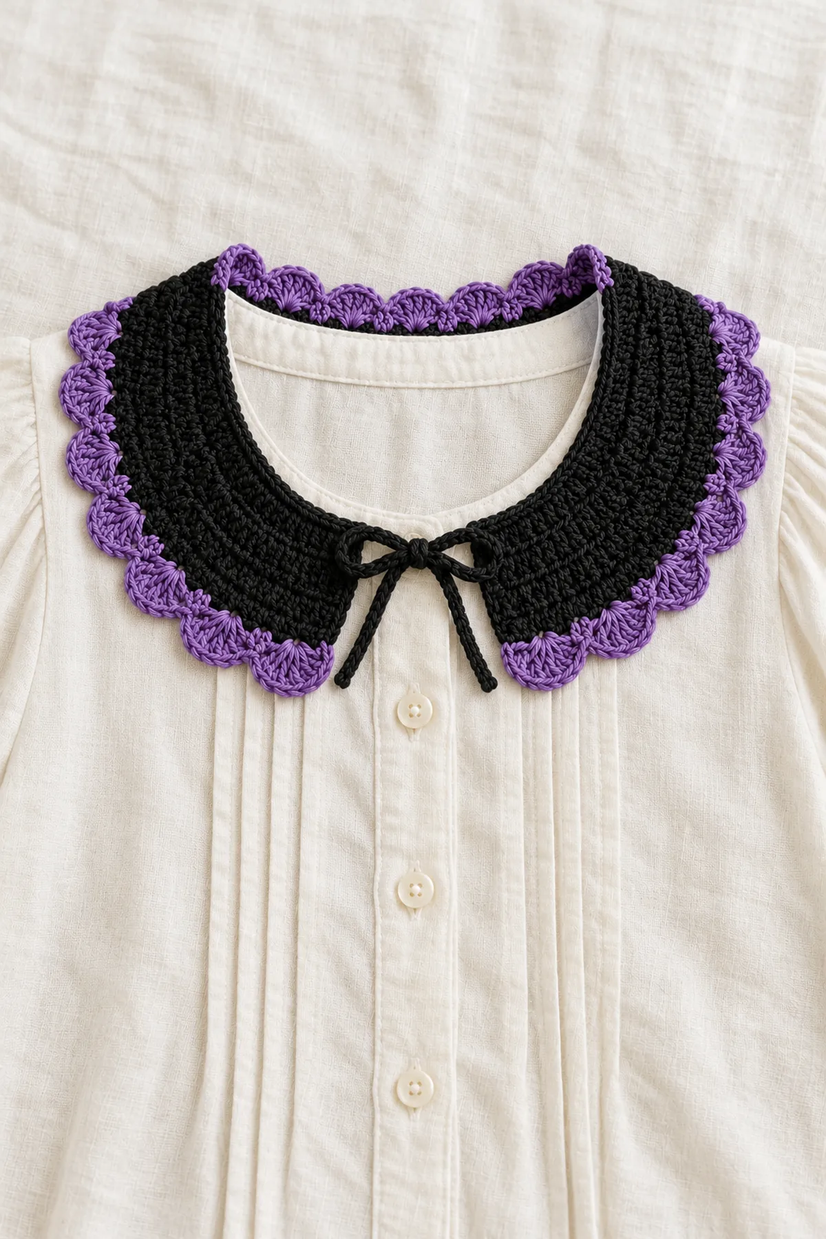 Same shallow black crochet crescent collar with violet fan scallops laid over neckline of an unworn cream blouse on a flat table, short black flat ties in a small loose front bow with an open gap. No person, mannequin neck, jewelry or long dangling cords