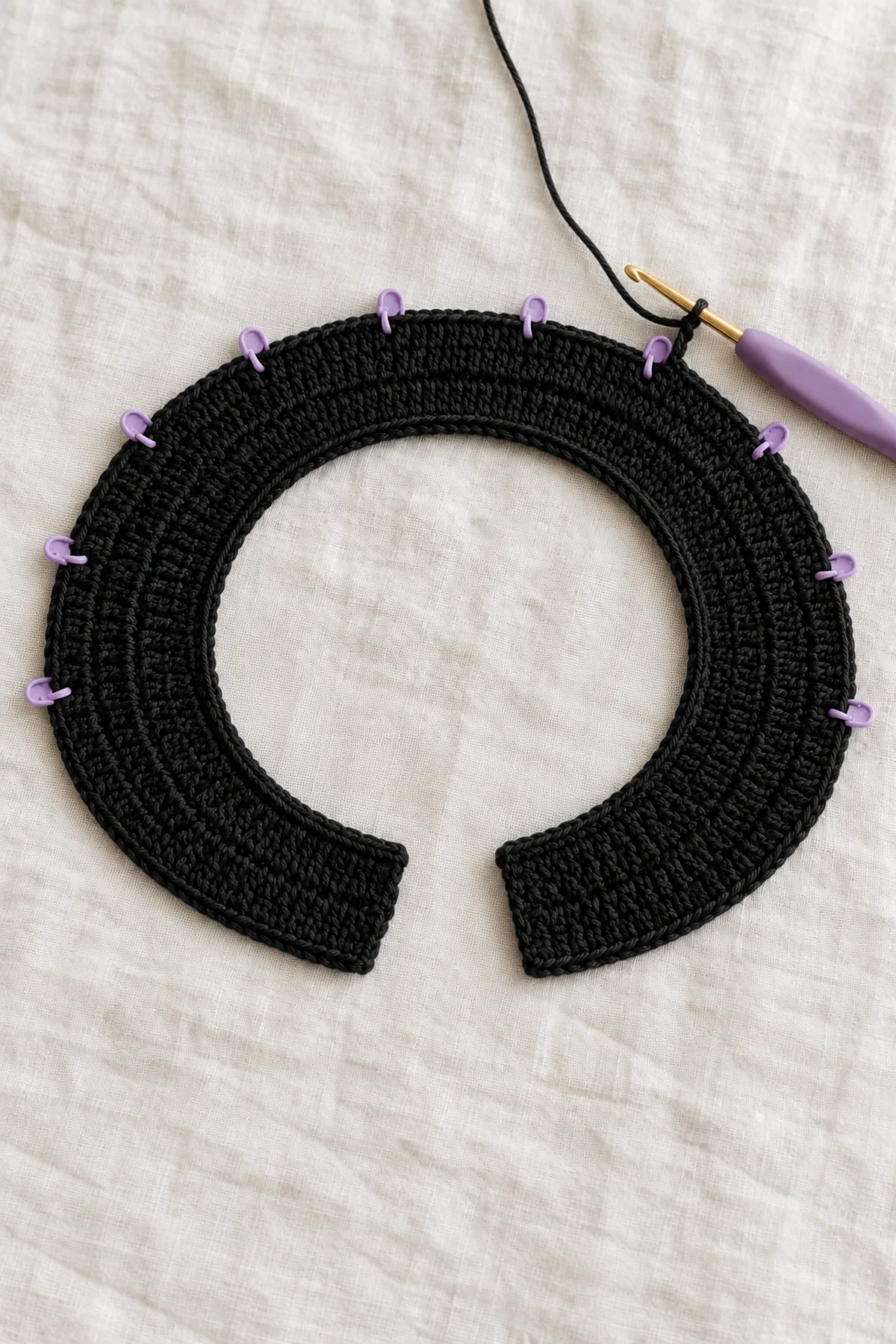 Black crochet neckline strip growing into a shallow curved band, first sc row and first two widening half-double-crochet rows complete, hook and evenly spaced markers on active edge; no violet fans, no ties
