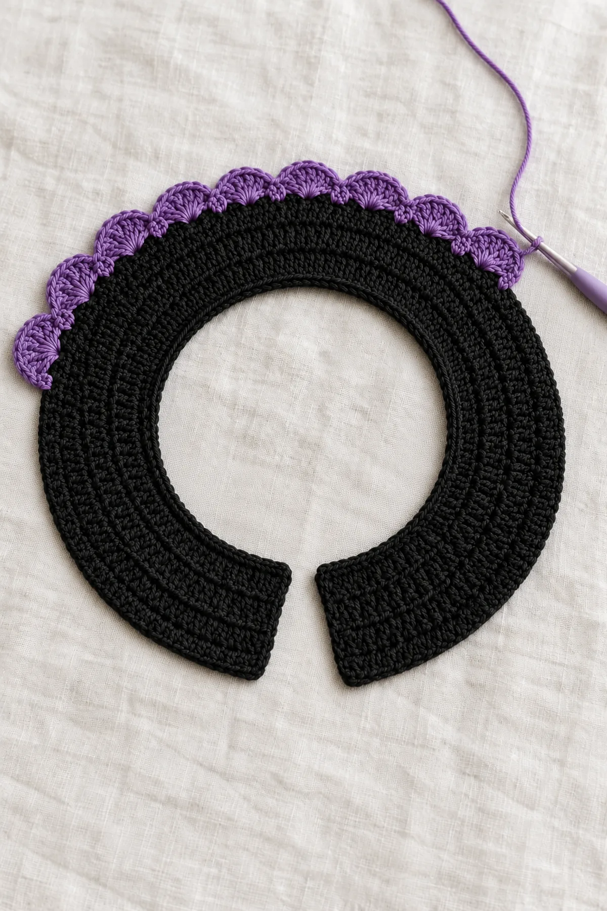 Flat curved black crochet collar body with all four expanded hdc rows complete and violet five-dc fan border partially crocheted along outer curve, hook at last completed fan, no ties yet
