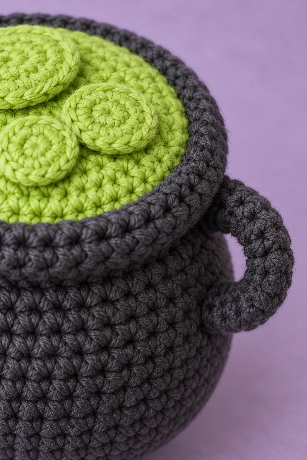 Macro side top view of same charcoal cauldron showing thick rolled crochet rim sewn over closed lime top seam, edges of flat sewn lime bubble discs and one flat curved charcoal side handle with securely stitched ends