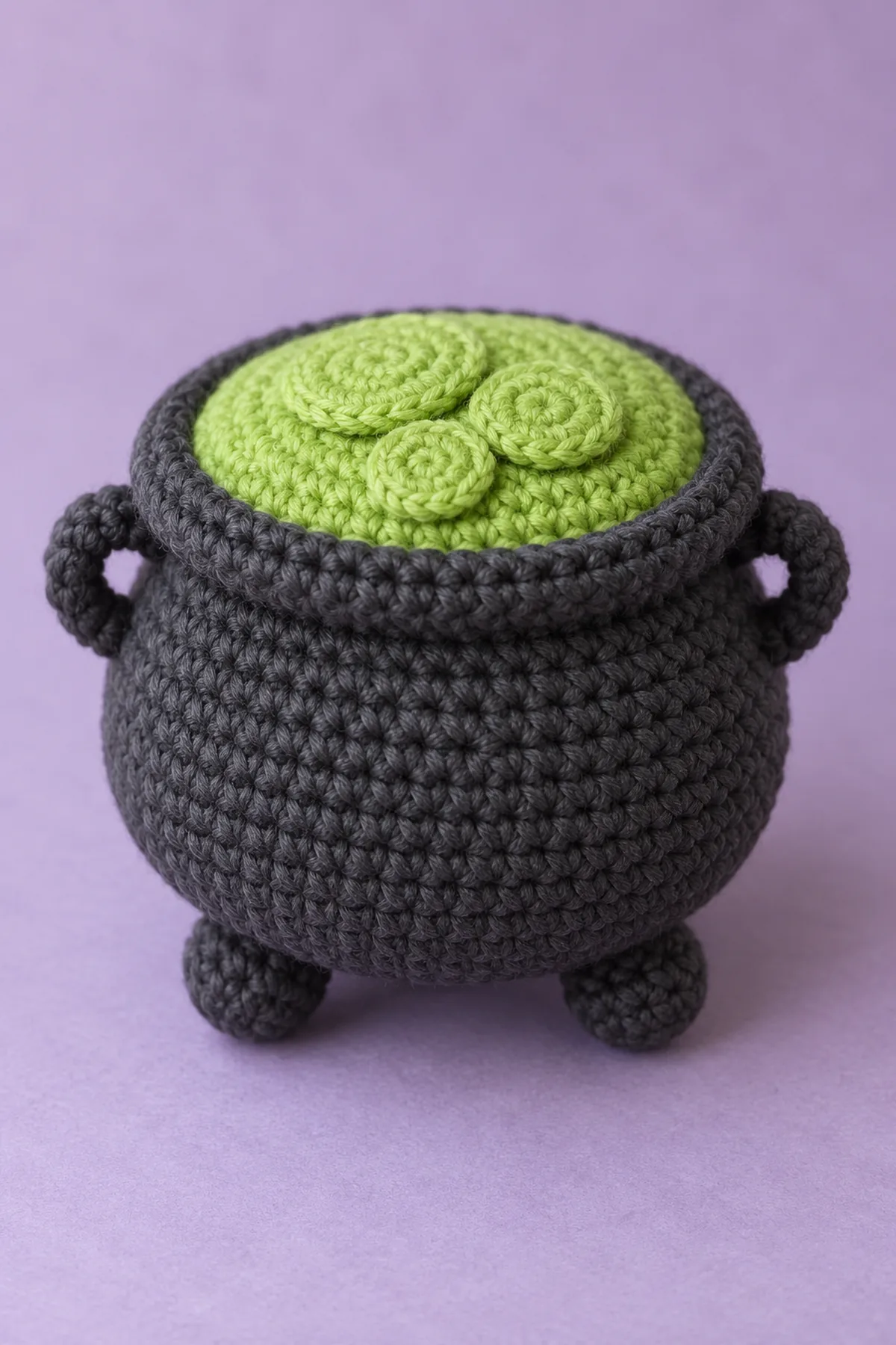 Original tiny closed stuffed charcoal cotton crochet cauldron, squat rounded pot belly about3.2in wide, THREE small rounded bobble feet integral underneath, thick rolled charcoal rim, closed flat lime top with THREE sewn flat overlapping lime circular bubble discs of graduated size, TWO small flat charcoal arch handles at opposite sides below rim. On lilac surface. No face, no liquid, no open hole, no floating balls, no bail handle or props