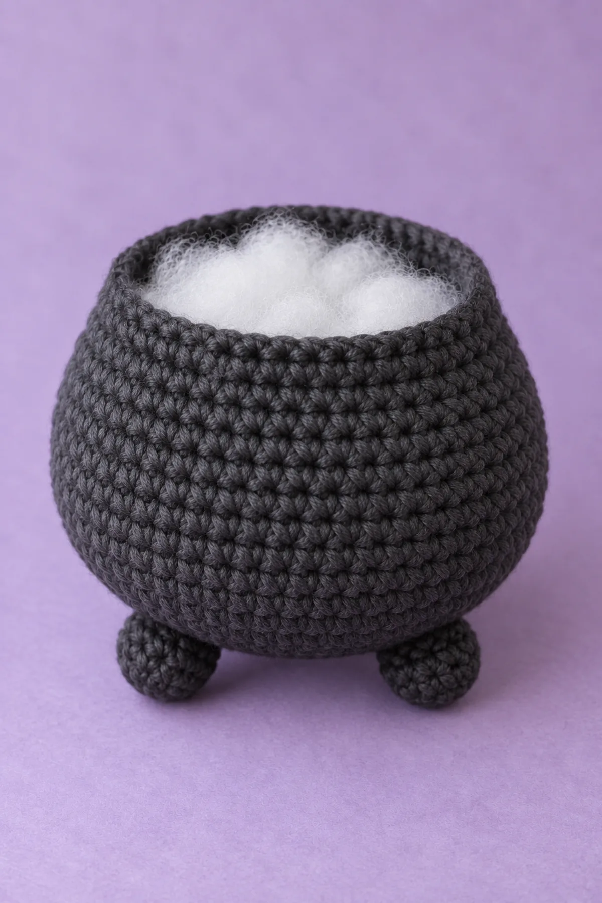 Unfinished charcoal crochet pot body sitting on three small integral bobble feet, rounded belly and open top with visible white stuffing inside, no lime top, no bubbles, no rolled rim, no handles yet